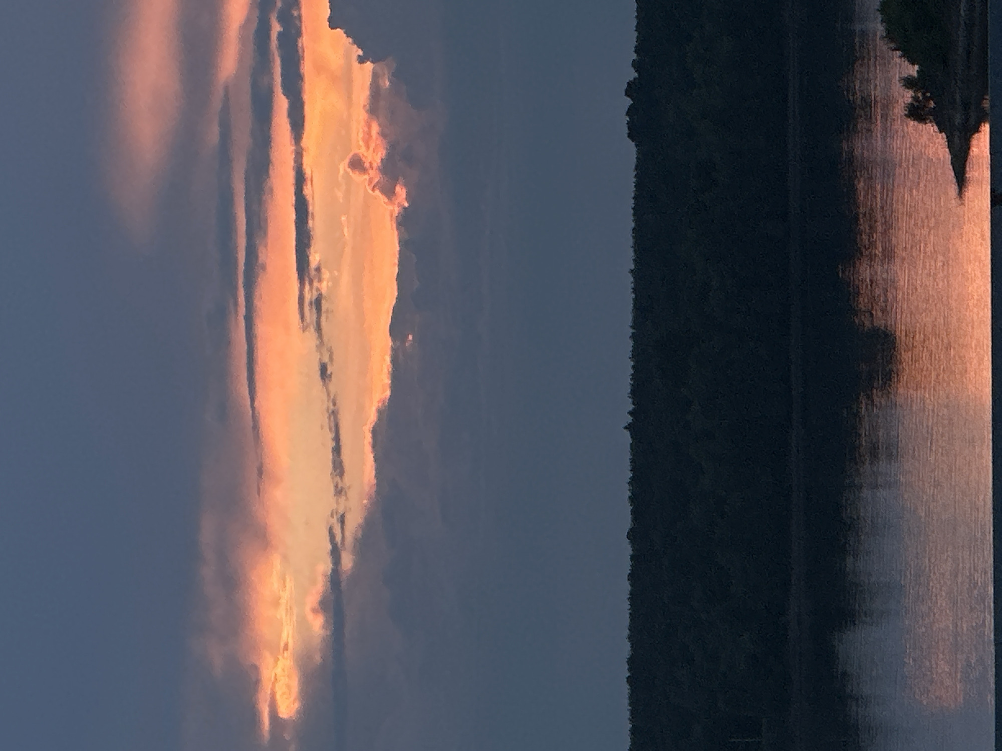 The image depicts a sunset over a body of water, likely a reservoir, on the outskirts of Chaikovsky, Russia. A dense line of dark green trees forms the horizon, separating the water from the sky. The sky is dramatic, with clouds in varying shades of deep blue and gray, illuminated from behind by the vibrant orange and pink hues of the setting sun. The light of the sunset is reflected in the still water, creating a shimmering orange and pink surface. The overall atmosphere is serene and natural. There are no people or man-made structures visible in the immediate scene.