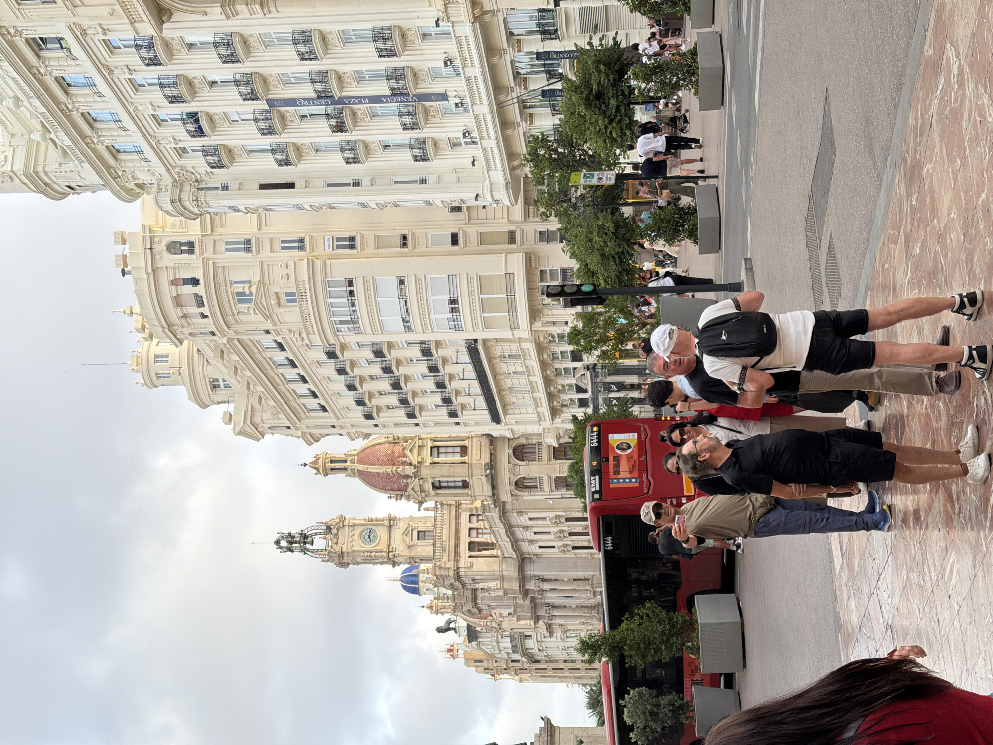 The image captures a street scene in a city, possibly Valencia, Spain, given the architectural style of the buildings and the presence of a red bus with markings that suggest public transportation in Europe. In the foreground, a group of people are gathered on a paved area with a marble-like pattern. One man in a light brown t-shirt and blue pants is holding up a phone, appearing to take a selfie. To his right, a man in a black polo shirt and black shorts is standing and looking to the side. Another man in a white t-shirt and black shorts with a backpack is facing away from the camera.
Behind the group, a bright red public bus is parked. The bus has various advertisements and route numbers visible on its side. In the background, several grand buildings with ornate facades and balconies line the street. The architecture features classical elements, with curved roofs and arched windows, suggesting historical significance. The sky is overcast, and trees are planted in planters along the street, contributing to the urban landscape. A traffic light is visible, indicating an intersection. The overall impression is that of a busy city square or main thoroughfare, with tourists and locals interacting amidst striking architecture.