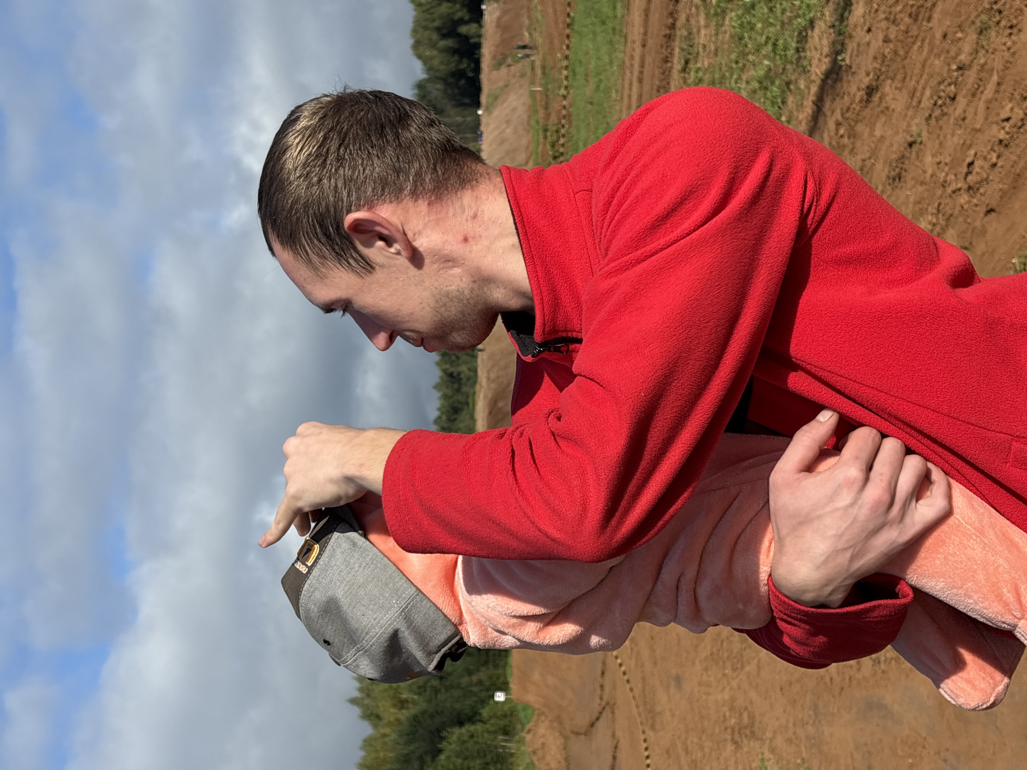 A man in a red jacket holds a child wearing a pink fuzzy outfit and a grey baseball cap. The man is looking upwards and pointing at something above, possibly the sky. The child is partially obscured by the man's arm. In the background, there is a dirt track with jumps, indicative of a motocross or similar racing venue. Green grass and trees are visible beyond the track, suggesting an outdoor, possibly rural, location. The sky is cloudy. The location is identified as Balakovo, Russia.