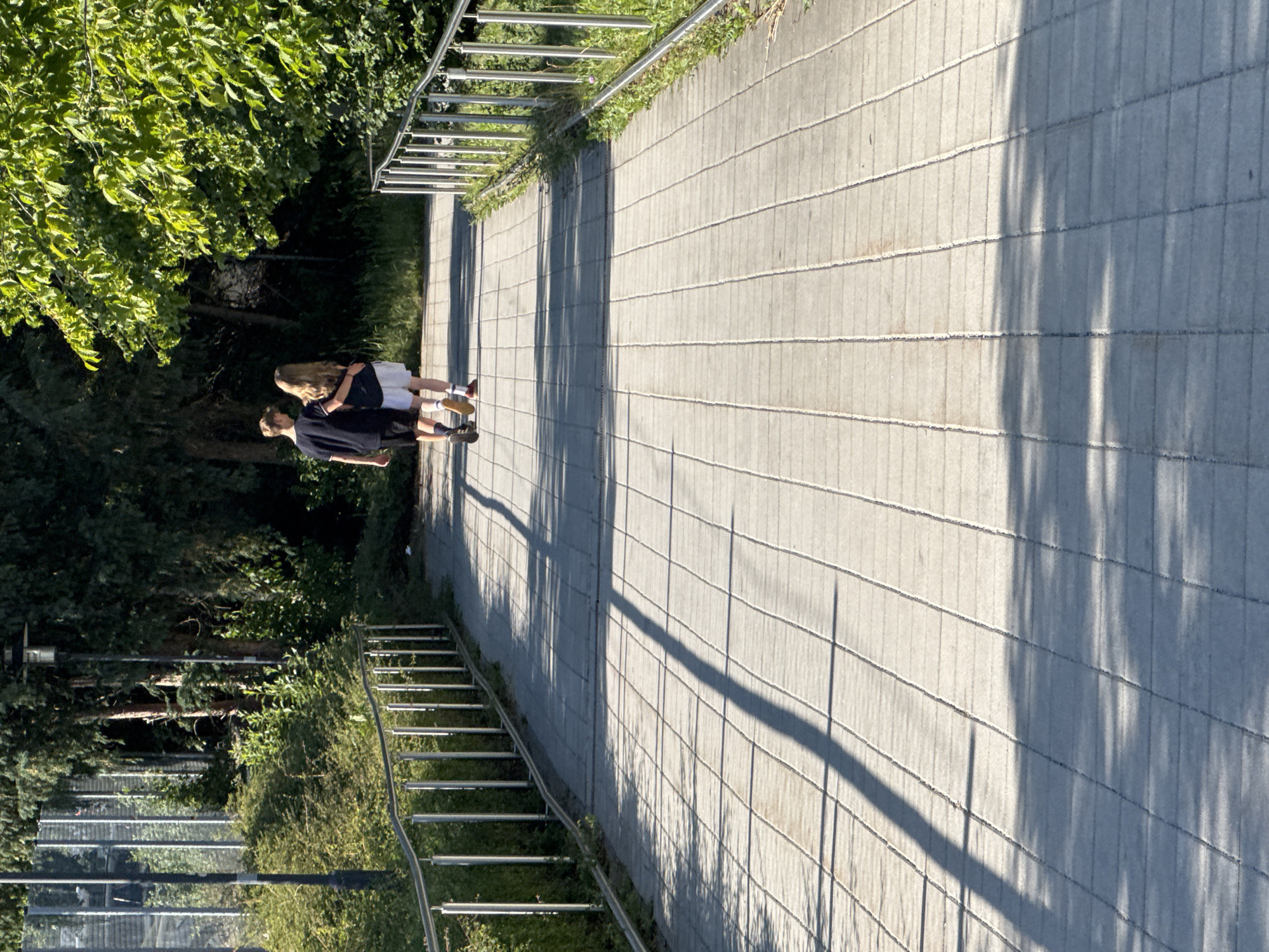 The image shows two people embracing on a paved walkway beside a lush green area. The walkway is made of rectangular concrete slabs laid in a grid pattern. The people are standing side-by-side, facing away from the camera, with the person on the left with blonde hair wrapped in the arms of the person on the right. Both are dressed casually, the person on the right in a dark t-shirt and shorts, and the person on the left in a white shirt and dark shorts. They appear to be in a relaxed and affectionate pose.
To the left of the walkway, there is dense green foliage from trees and bushes, with some visible sunlight filtering through. A metal railing runs along the edge of the walkway, separating it from the greenery. To the right of the walkway, the concrete continues, with long, distinct shadows of trees or other structures cast across it, suggesting it is a sunny day, likely in the late afternoon or early evening due to the length of the shadows.
In the background, there are glimpses of a metal staircase leading down into a grassy area, and more trees. The overall scene suggests a pleasant outdoor setting, possibly a park or a public path. No text is visible in the image. The location is identified as Höhenkirchen-Siegertsbrunn, Germany.