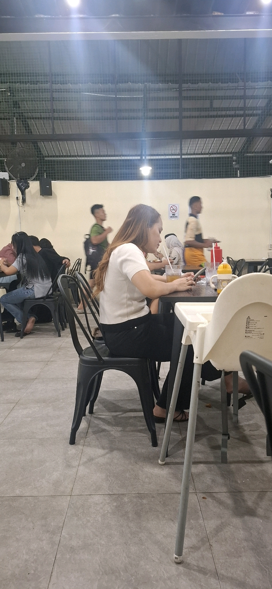 The image depicts a casual dining area, possibly a food court or an outdoor seating section of a restaurant. In the foreground, a woman with long, brown hair is seated at a dark table, looking down at her phone. She is wearing a white short-sleeved shirt and black pants. Her right hand is holding a straw in a glass of amber-colored liquid. To her right, there is a baby high chair and another person partially visible. The floor is tiled with a light gray pattern. In the background, several other individuals are seated at tables or walking. One man in a green shirt and backpack is seen walking, and another man in a yellow and orange outfit is also walking. A "No Smoking" sign is visible on the wall behind them. The ceiling structure appears to be industrial, with exposed metal beams and a grid-like material overhead, suggesting an open or semi-open space. A large black fan is mounted on the wall on the left side of the image. The overall lighting suggests an evening or dimly lit indoor setting, with bright lights contributing to the glare. The context provided by the user indicates this scene is located in Surakarta, Indonesia.