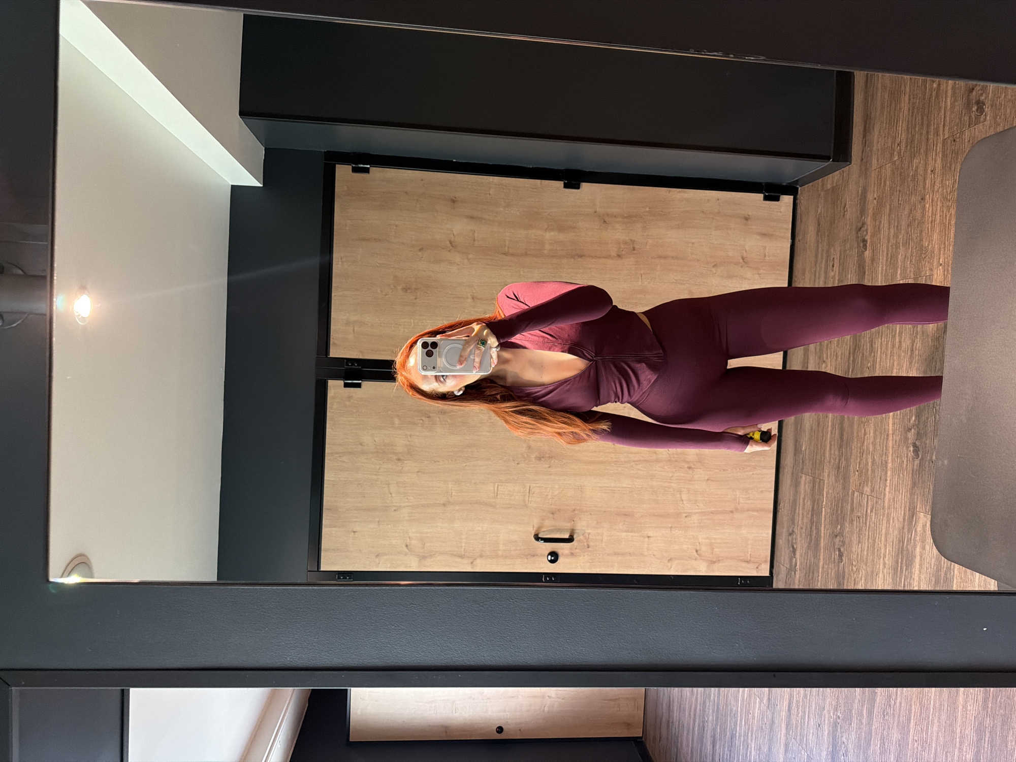 A full-length, rotated mirror selfie captures a woman in a maroon-colored activewear set. She holds a smartphone in her right hand, partially obscuring her face. The scene appears to be within a room with wooden paneled walls, dark gray accents, and wooden flooring. A white light fixture is visible in the upper left of the mirror's reflection. The location's connection to Paris, France, is not discernible from the visual elements in the image.
