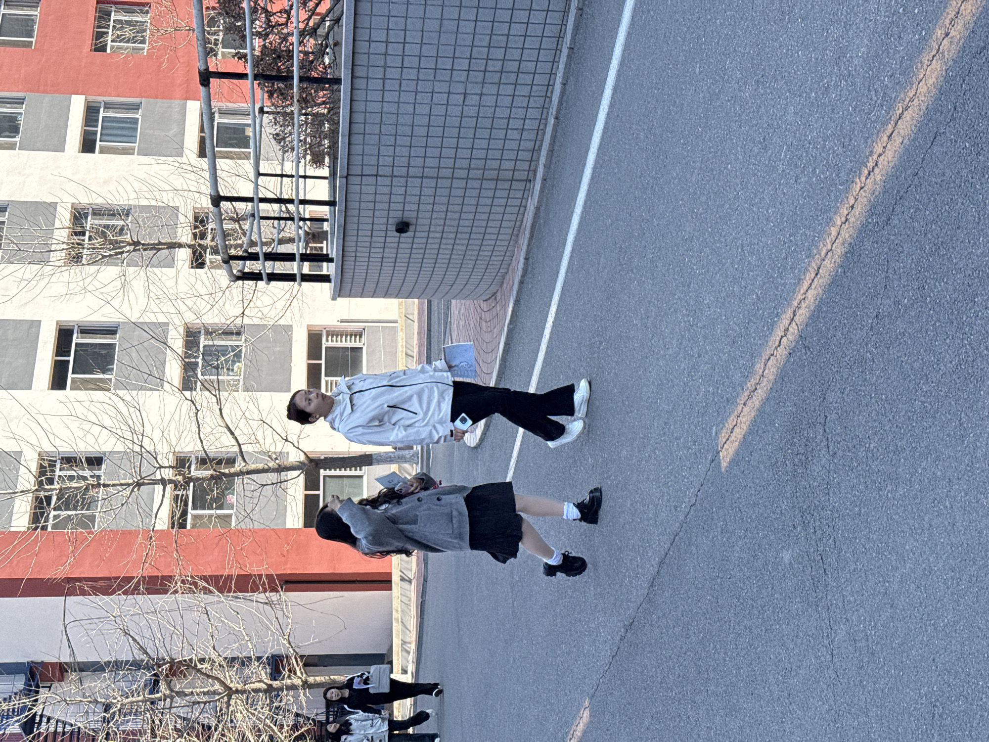 The image captures a scene in Zhongtun, China. Two individuals, one wearing a light-colored jacket and dark pants, and the other in a gray jacket and dark skirt, stand on an asphalt surface adjacent to a building with red and gray walls and many windows. A bare tree stands nearby. Two more people walk further away in the same direction.