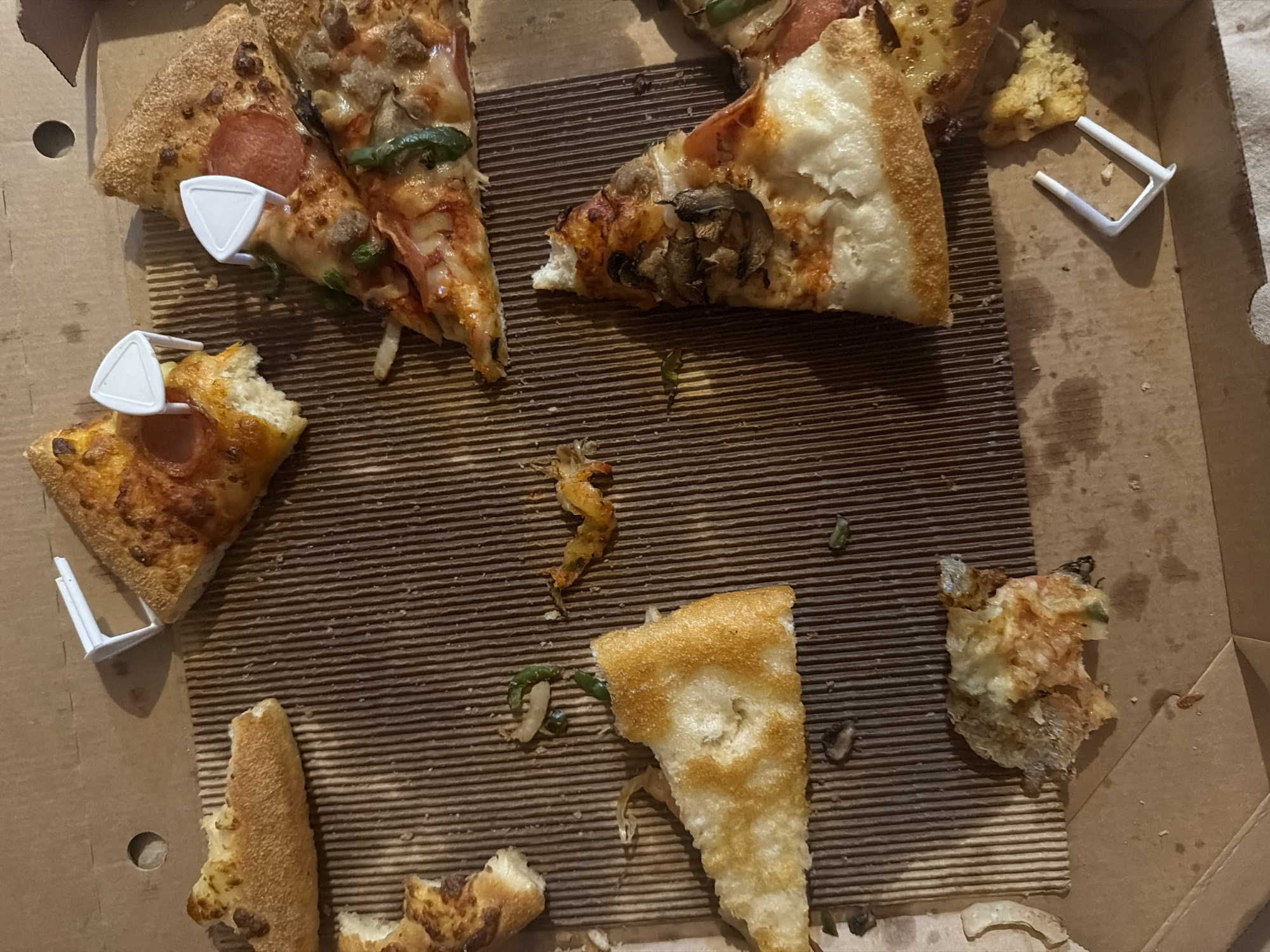 The image shows a pizza box with several slices of pizza, some of which have been partially eaten. There are also pizza saver inserts scattered throughout the box. The pizza appears to be a variety of toppings, including pepperoni, mushrooms, and green peppers. The pizza box is made of cardboard and has a corrugated interior. The scene is a close-up, overhead shot of the pizza box, and it is difficult to determine the exact location within Tersefanou, Cyprus. However, based on the presence of a pizza box, it is likely a residential or commercial setting where pizza is consumed. There are no people visible in the image, and no actions or interactions are depicted. The environment is limited to the interior of the pizza box.