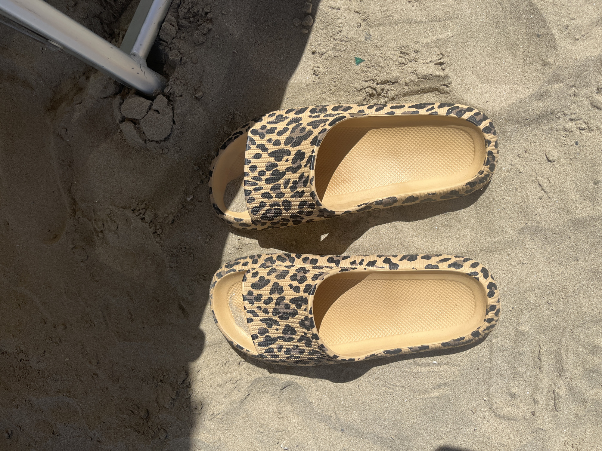 The image shows two leopard-print slides on a sandy surface, likely a beach. The slides are tan with black spots and appear to be made of a soft, possibly rubbery, material. The sand is textured, with visible grains. In the upper left corner, there's a section of a metallic object, possibly part of a chair or beach furniture. The lighting suggests it is daytime. The location is Unknown, Unknown.