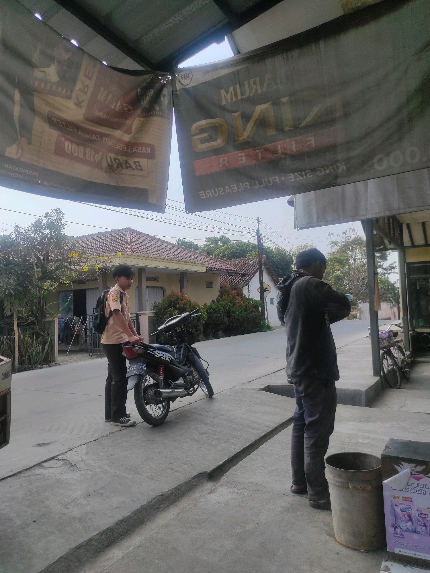 The image depicts an outdoor scene in Ngamprah, Indonesia. In the foreground, a young male wearing a school uniform with a backpack is standing next to a parked motorcycle. He is looking towards the motorcycle. To the right, another male wearing a dark jacket and pants is standing and looking at his phone. A large trash bin and a box of laundry detergent are visible near him. In the background, a residential house with a tiled roof and a yard with trees and bushes is seen. Several banners are hanging overhead. The scene appears to be on a street or sidewalk.