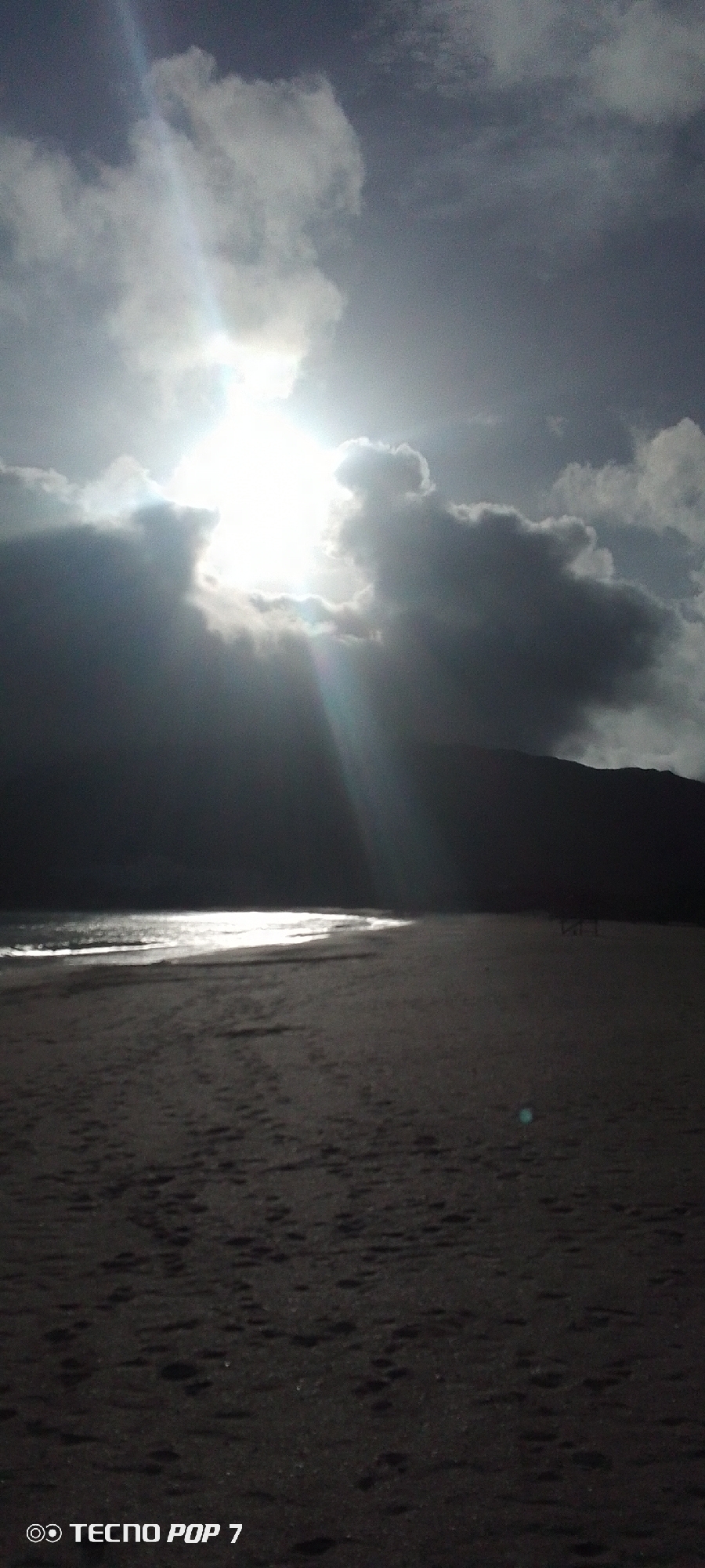 The photograph shows a beach scene near Pedro González, Venezuela. The sky is overcast with the sun breaking through a gap in the clouds, creating a bright glare and beams of light. A dark silhouette of a mountain range is visible in the background. The beach stretches from the lower edge of the frame towards the water, which reflects the sunlight. Footprints mark the sandy surface. The words "TECNO POP 7" and two concentric rings are visible in the lower left corner of the image. There is nobody in the photograph.