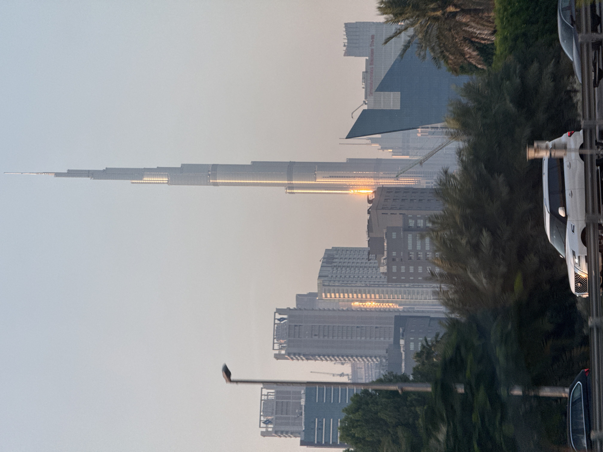 The image shows a view of the Dubai skyline, with the Burj Khalifa as the dominant feature. The skyline is partly obscured by trees and other foliage in the foreground. Several other high-rise buildings are visible, along with the sun reflecting off their surfaces. A street lamp is also visible in the foreground, as well as parts of cars parked along the side of the road. The sky is light grey and hazy.