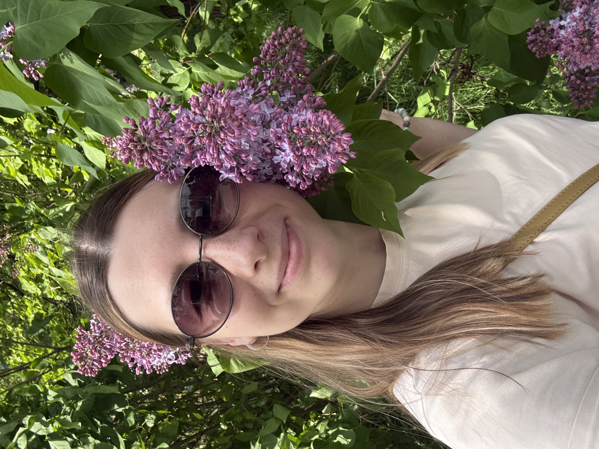A woman with light brown hair is wearing sunglasses and lying amongst lilac bushes. The woman is wearing a light-colored shirt with a strap across her shoulder. The location is not discernible as Moscow, Russia.