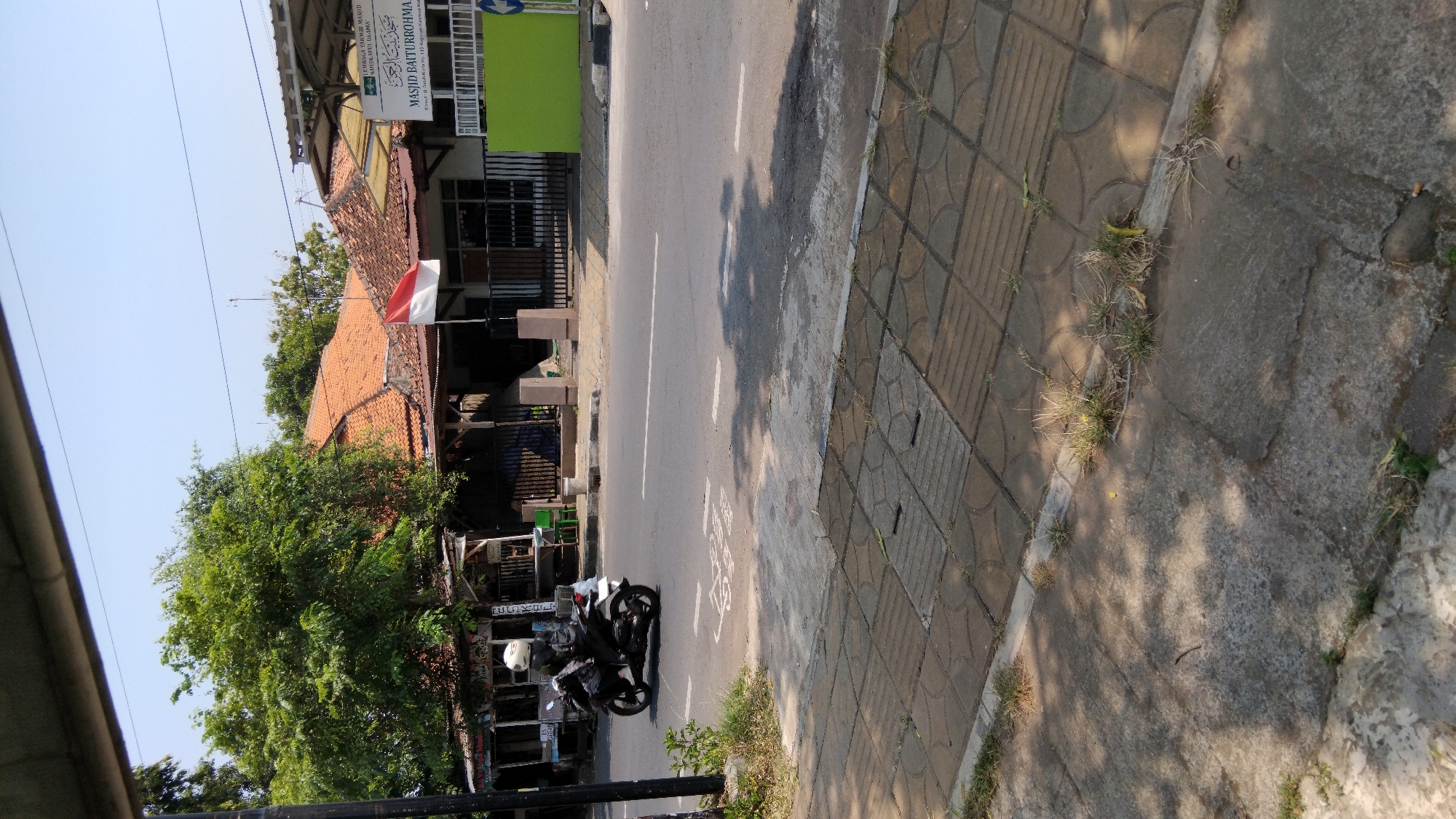 The image captures a street scene in Proyonangg, Indonesia, under a clear sky. In the foreground, a tiled pedestrian walkway runs along the side of a paved road. The road itself features white lane markings and the shadow of trees. A person wearing a white helmet rides a black scooter on the road, moving from left to right.
In the background, across the road, are several buildings. The most prominent is a building with an orange tiled roof and a white facade, identified by a sign as "MASJID BAITURROHMA" and associated with "LEMBAGA TAKMIR MASJID NAHDLATUL ULAMA." Next to it, a bright green wall stands out. To the left of these buildings, there's a tree with dense green foliage, and further left, a small roadside stall or structure with a weathered roof is visible. A red and white Indonesian flag is prominently displayed on a pole in front of the mosque. The environment appears to be a typical urban or semi-urban area, likely a residential or commercial district.