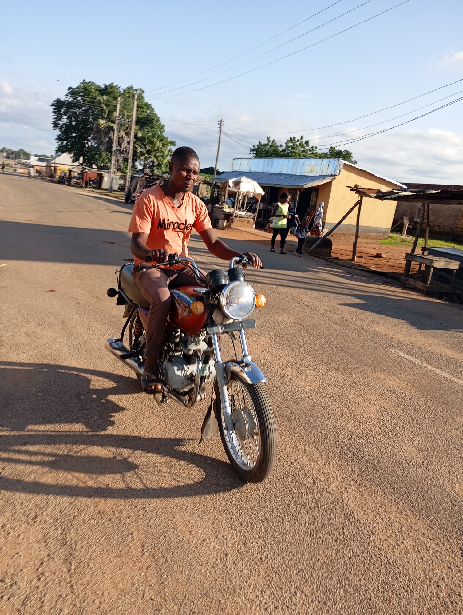 A young man is riding a motorcycle on a paved road in what appears to be a rural or semi-urban area of Shendam, Nigeria. He is wearing an orange t-shirt with the word "Miracle" printed on it and brown trousers. The motorcycle is orange and silver with a visible engine and exhaust. The sun is casting long shadows from the man, motorcycle, and other objects in the scene, indicating it is either early morning or late afternoon. In the background, there are several small buildings with corrugated metal roofs, some appearing to be shops or market stalls. A few other people are visible in the background near these structures. Trees and power lines are also present, typical of a developing area. The road surface is asphalt with some gravel. The sky is clear and blue with some faint clouds.