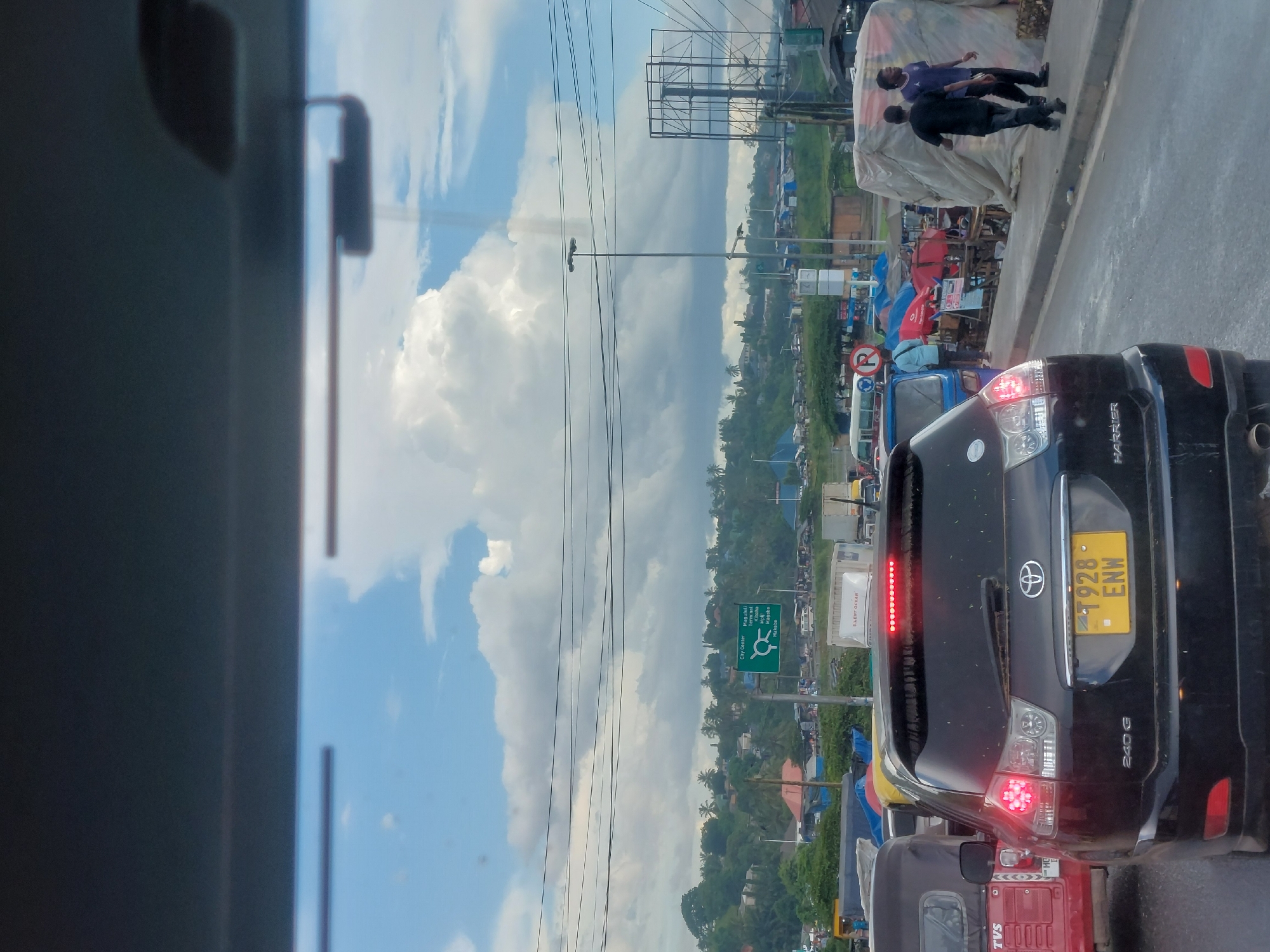 The image depicts a traffic jam in Dar es-Salaam, Tanzania. The camera is positioned inside a vehicle, looking out at other vehicles and roadside activity. A black Toyota Harrier with license plate T928 ENW is the primary vehicle in view. The street is lined with various stalls or informal businesses. Two men are standing on the curb, near some goods covered with white cloth. A green road sign indicates directions to the City Center, Magufuli Terminal, Kibaha, Magohe, and Makabe. A red "Silent Ocean" truck is seen in the traffic. Above the scene is a partially cloudy sky. Power lines traverse the view.