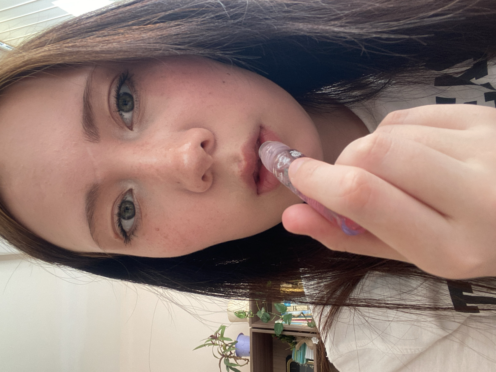 A young woman with brown hair is applying a lip gloss. She has fair skin, freckles and green eyes. She is wearing a white shirt. In the background is a bookcase, plants, and a white wall. The location is unknown.
