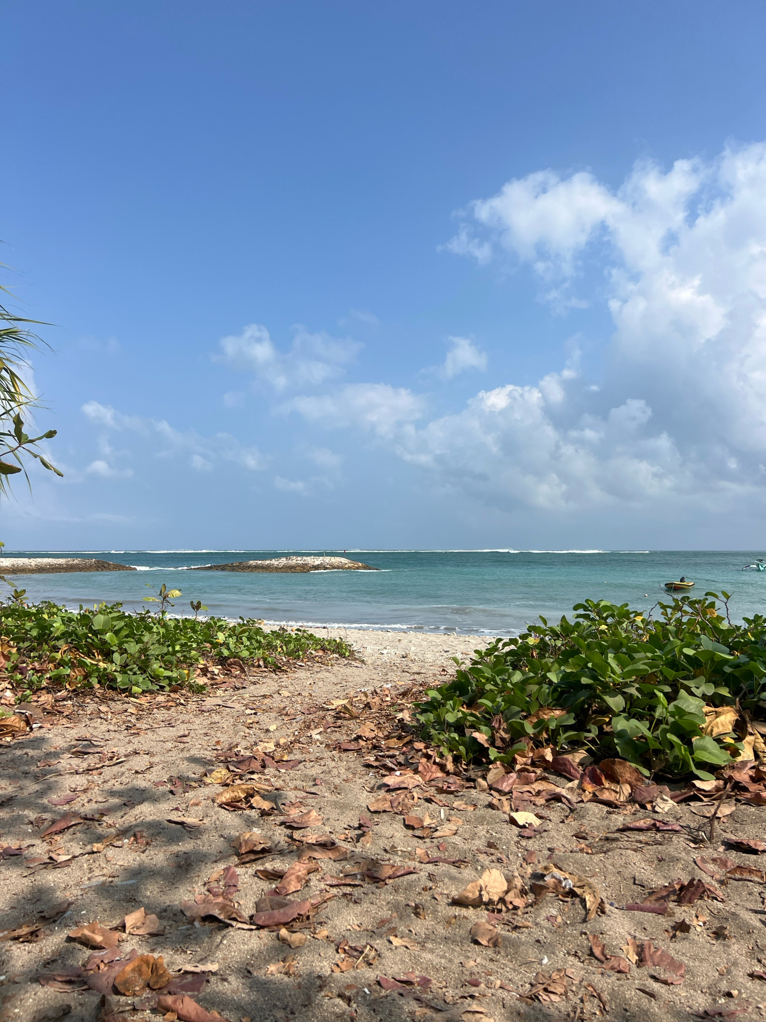 The image depicts a serene beach scene in Kuta, Indonesia. In the foreground, a sandy path is strewn with fallen brown leaves, bordered by lush green vegetation on either side. Beyond the vegetation, the turquoise ocean stretches towards the horizon, with gentle waves breaking along the shore. Two man-made rock structures, likely breakwaters, jut out into the water, with a distant person standing on one of them. A small yellow boat is visible on the right side of the frame, anchored in the sea. The sky above is a clear blue, dotted with fluffy white clouds. The overall atmosphere is peaceful and natural.