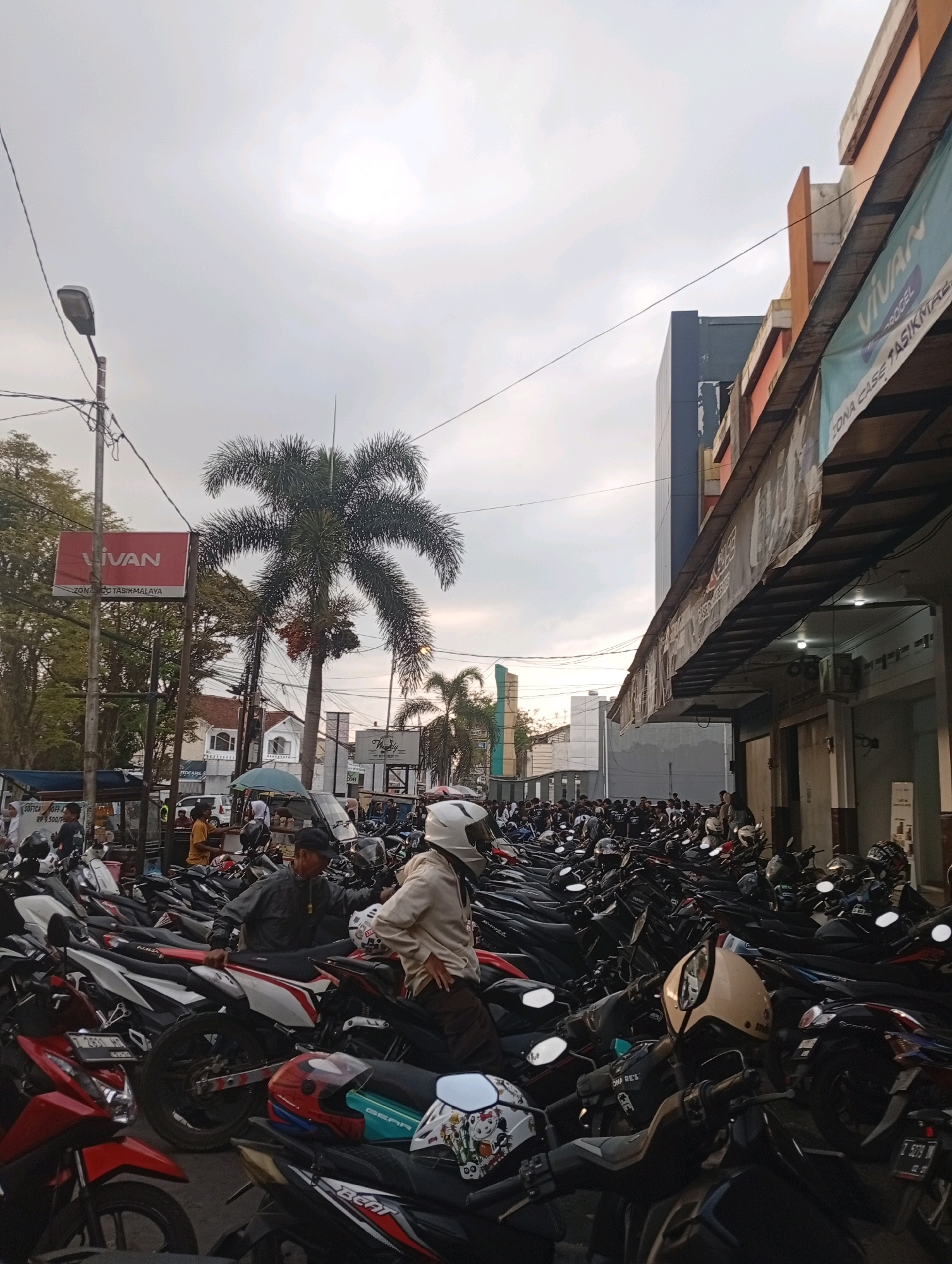 This is a street-level, eye-level, full-frame, vertical shot of a crowded motorcycle parking lot in Tasikmalaya, Indonesia. The sky is overcast with clouds. In the foreground and midground, hundreds of motorcycles are parked in rows. The motorcycles vary in color, with black, red, white, and silver being prominent. Some motorcycles have helmets on them.
In the midground, several people are visible. Two men, one wearing a black jacket and a cap, and another wearing a beige shirt and a white helmet, are standing near motorcycles. Other individuals are scattered among the parked vehicles, some appearing to be pedestrians, others possibly owners of the motorcycles.
In the background, there is a building with an orange and white facade and a blue and grey modern building behind it. A banner with the word "VIVAN" is visible on the orange building, and another banner with "ZONA CASE TASIKMALAYA" is also present, indicating the location. A palm tree stands tall in the middle ground, adding a tropical element to the urban scene. Several streetlights and power lines crisscross the sky. The overall impression is a busy urban environment with a significant focus on motorcycle transportation.