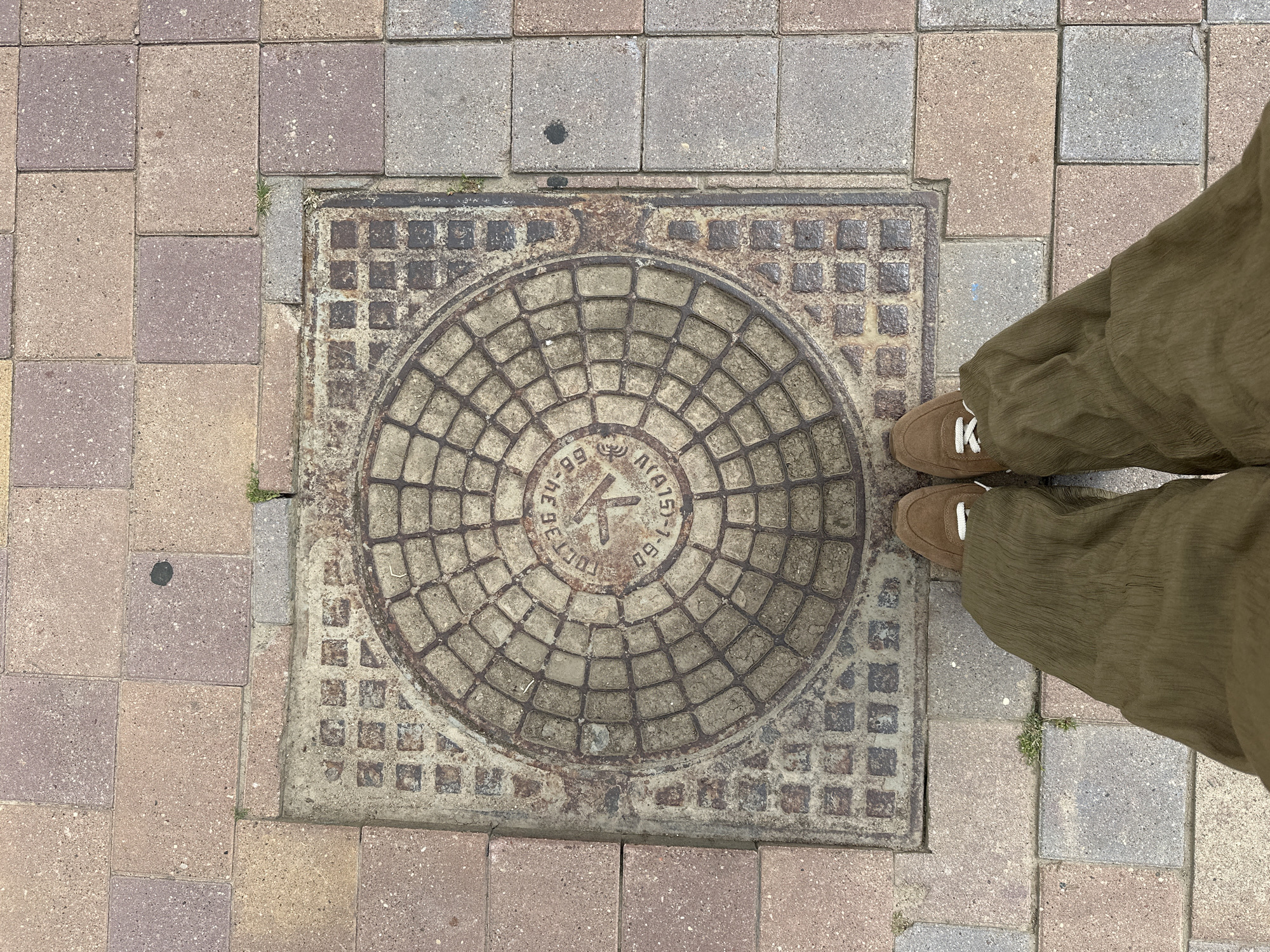 This is a top-down, eye-level, medium shot of a person standing on a sidewalk in Minsk, Belarus. The person is wearing loose, olive-green trousers and brown suede sneakers with white laces. They are standing on a section of sidewalk composed of rectangular paving stones in varying shades of pink, beige, and gray. In the center of the frame is a square, cast-iron manhole cover set into the pavement. The manhole cover has a circular, patterned design in its center, with concentric rings and radiating lines. The outer edge of the manhole cover is embedded in a concrete frame, and around this frame, the paving stones are slightly uneven. The overall impression is of an urban environment with ordinary street furniture.