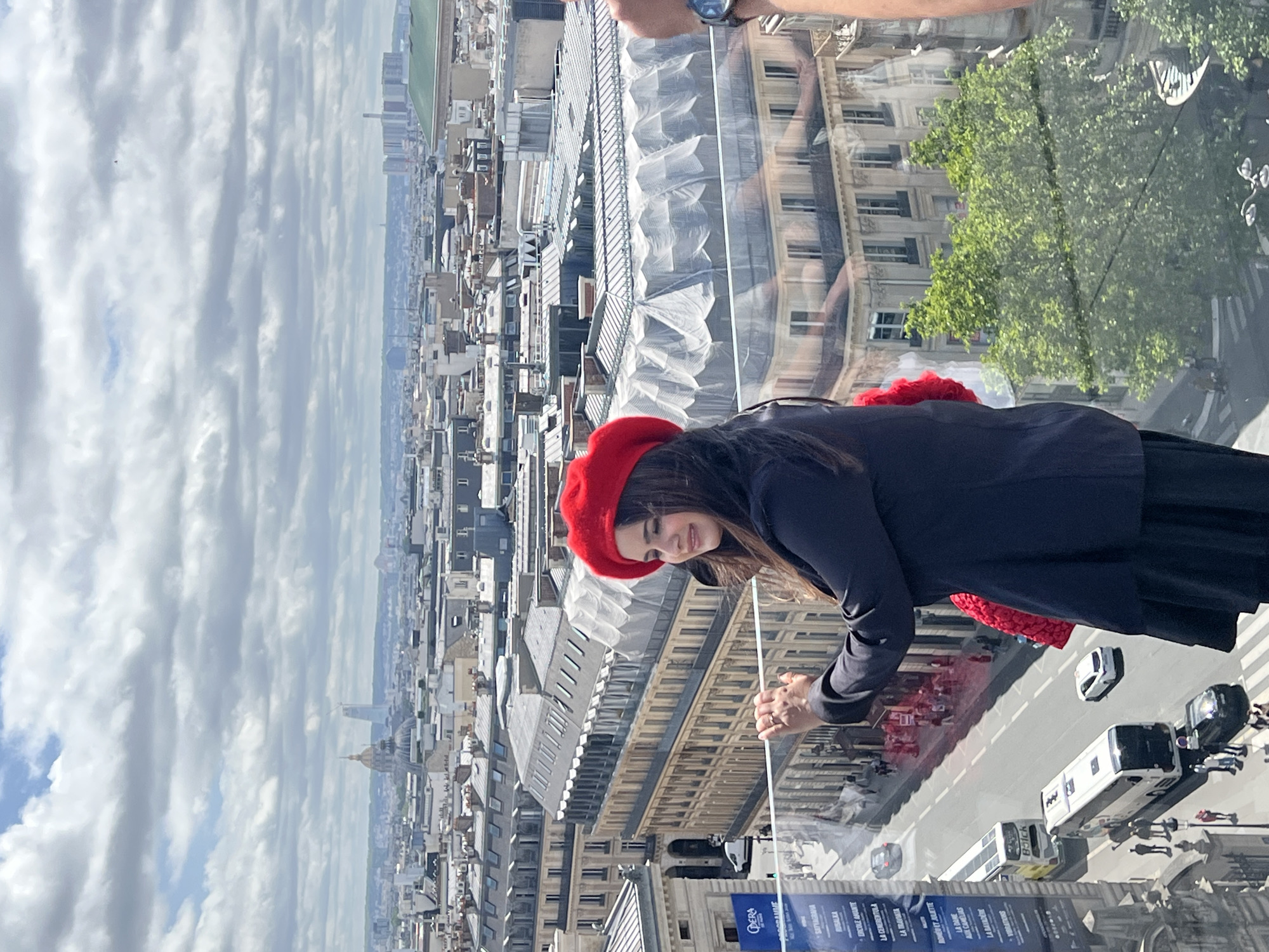 A woman wearing a red beret and a dark blue jacket stands on a balcony overlooking a city skyline. She is looking to her left and smiling. In the background, the cityscape of Paris, France, is visible with numerous buildings and a cloudy sky. A street with traffic, including buses and cars, runs below the balcony. The woman is holding onto a clear glass railing with her left hand. A red scarf or accessory is visible on her right side. To the right of the woman, part of a person's arm with a watch is visible. The image appears to be taken on a bright day, possibly during the daytime.