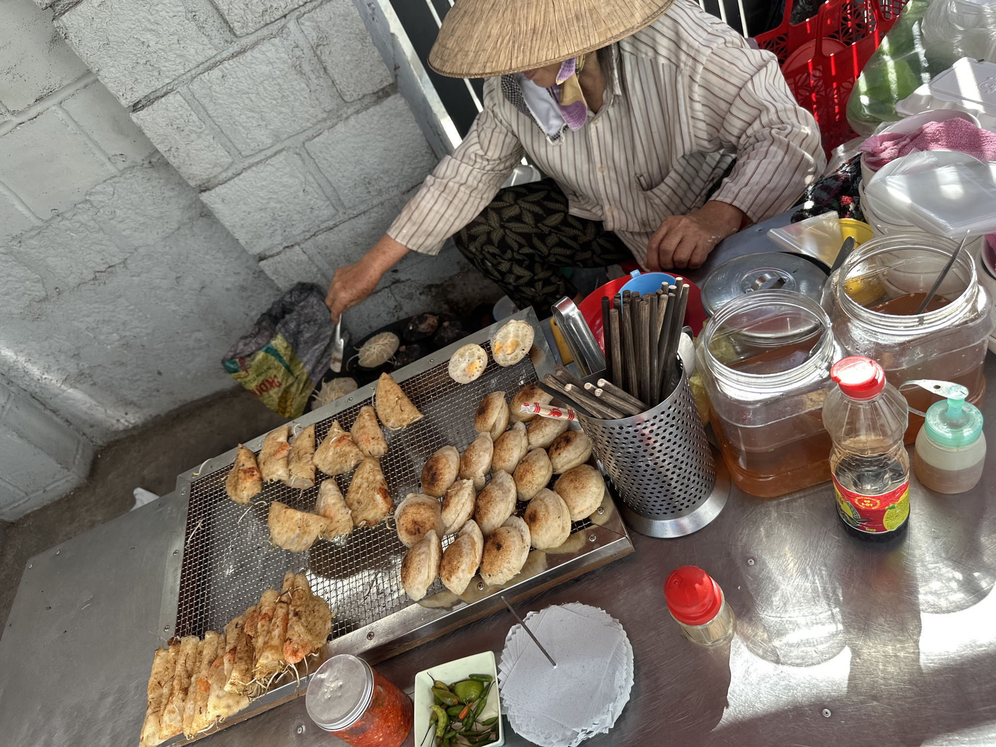 The image shows a street vendor in Bac Nha Trang Ward, Vietnam, preparing and selling food. The vendor, wearing a traditional conical hat, a patterned face covering, and a striped shirt, crouches beside a low table laden with food and cooking implements. She is using a spatula to manipulate small, round items on a cooking surface to her left.
On the table, there is a metal rack holding an assortment of prepared items: triangular-shaped snacks, round patties, and elongated bundles. A metal container filled with chopsticks and various condiments in clear jars and bottles are clustered nearby. Additional items are stacked in the background, including plastic containers and a red plastic crate. The background shows a gray brick or concrete wall. The scene is lit by natural daylight.