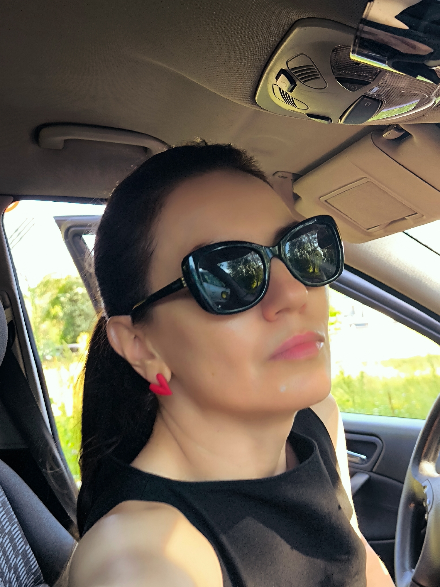 The image shows a woman wearing large black sunglasses and a black top, sitting in the driver's seat of a car. She has dark hair pulled back into a ponytail and is wearing bright pink heart-shaped earrings. The interior of the car is visible, including the ceiling, a sun visor, and part of the steering wheel. The outdoor scenery, with trees and sunlight, is visible through the car window. There is no discernible information in the image that indicates the location is the city of Riga, Latvia.