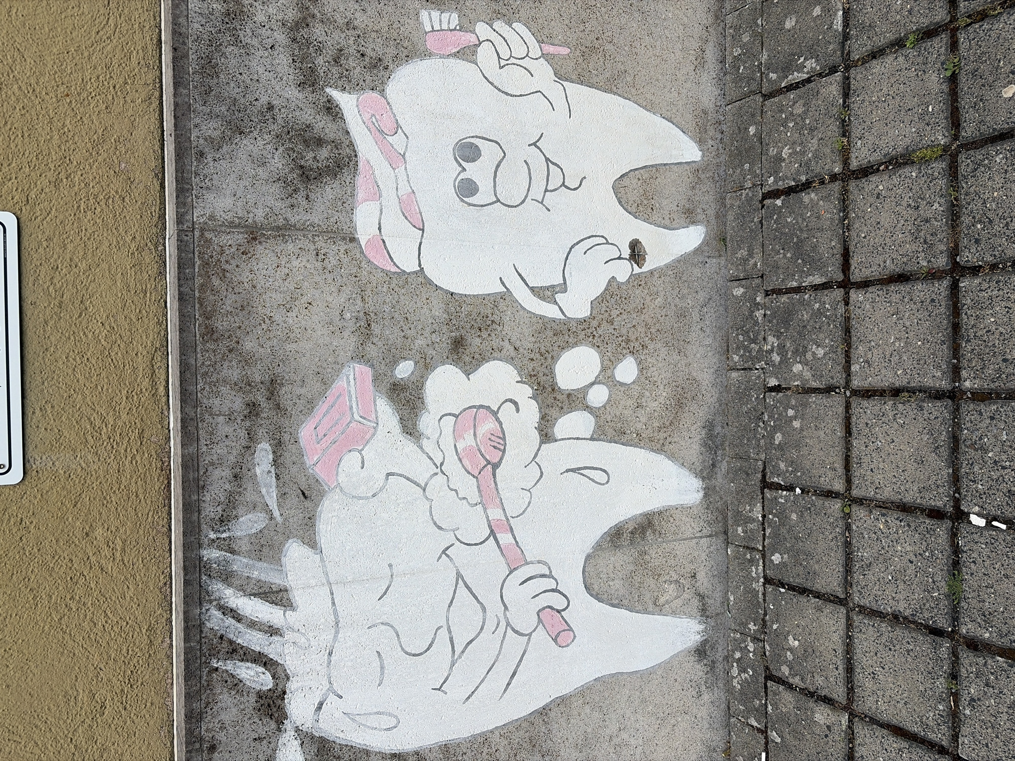 This is a photograph of a mural on a concrete wall in Würzburg, Germany, depicting two anthropomorphic teeth. The left tooth is brushing its teeth with a toothbrush and toothpaste, with bubbles and water splashes around it. The right tooth is also brushing its teeth with a toothbrush and toothpaste. The wall is textured and yellow above the mural, and the mural is painted on a grey concrete surface. Below the wall are grey cobblestones with some grass growing in the gaps.