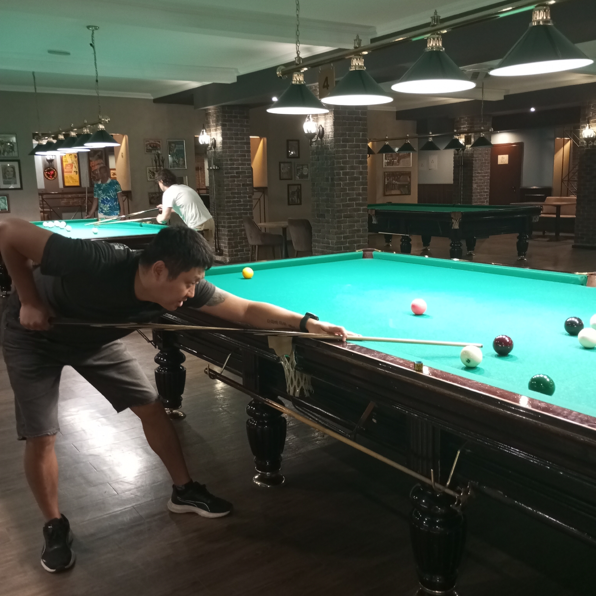 A man is playing pool in a dimly lit billiard room. He is wearing a black t-shirt and grey shorts and is in the process of striking a billiard ball with his cue. There are several other billiard balls on the green felt table. In the background, another person is visible near a second billiard table. The room has a rustic ambiance with exposed brick walls and overhead lighting fixtures. The specific location in Izhevsk, Russia, is not determinable from the image.