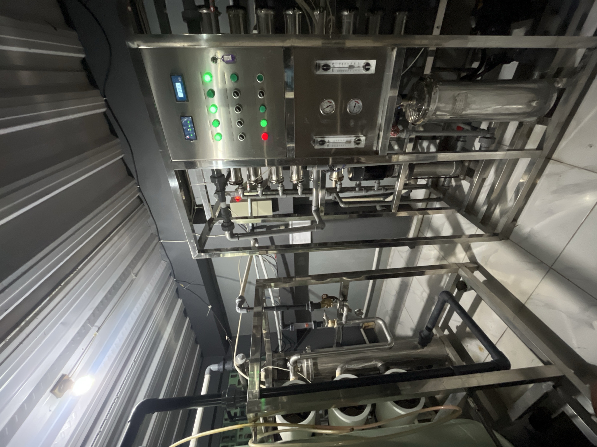The image depicts a water purification system, likely an industrial reverse osmosis unit, located indoors. The system is constructed primarily of stainless steel tubing, valves, pressure gauges, and filtration housings. A control panel with multiple indicator lights, some illuminated green and one red, is visible, along with digital displays. The ceiling appears to be corrugated metal with exposed lighting fixtures. The floor is tiled. The overall impression is one of an operational water treatment facility. The context of Pekalongan, Indonesia is not visually evident within the image itself.