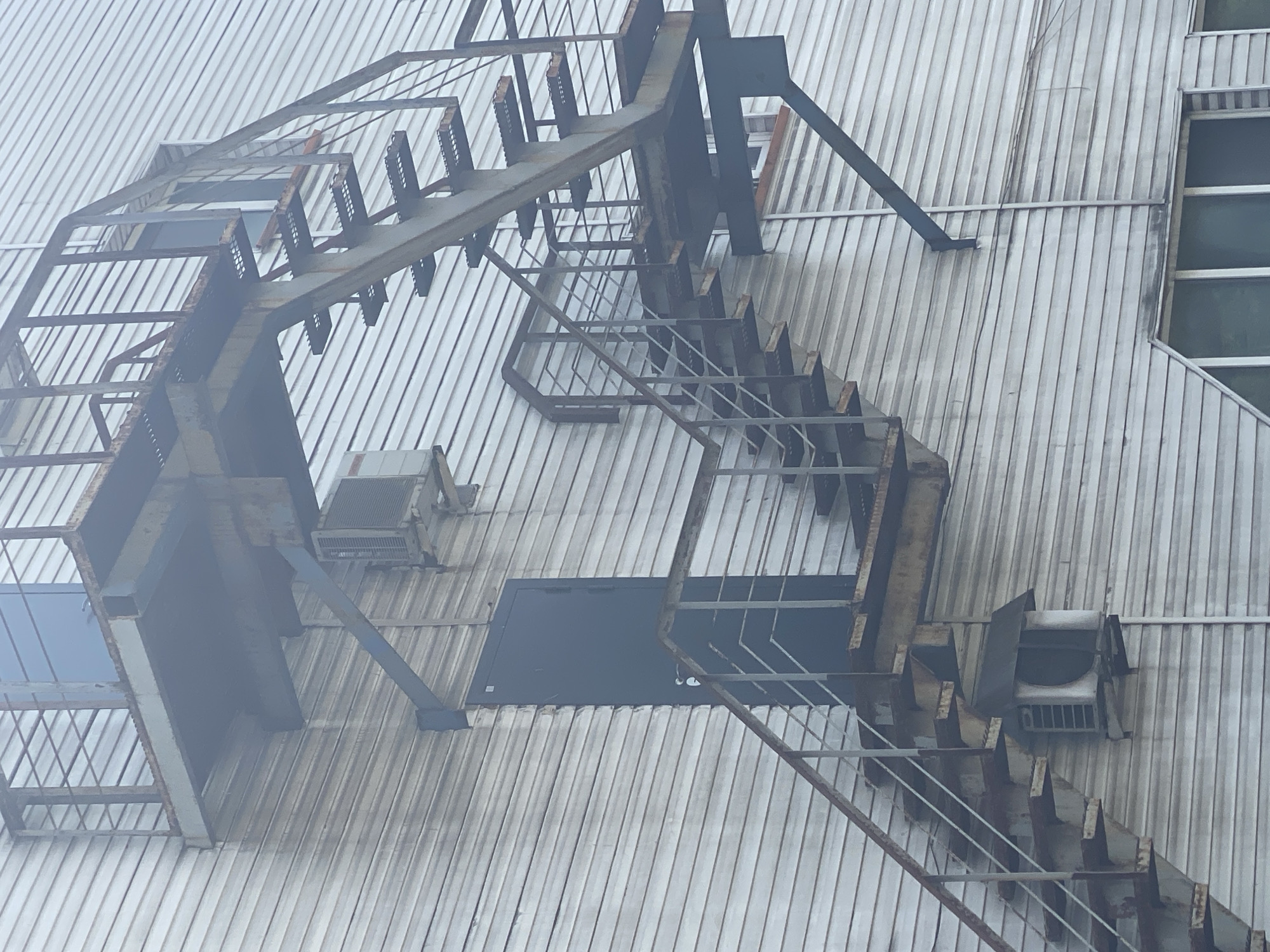 The image shows an outdoor metal fire escape staircase attached to the side of a building with horizontal siding. The staircase is weathered and appears to be made of rusted metal. There is a dark-colored door visible on the building wall, positioned below the upper landing of the staircase. Two window-like structures are also visible on the upper landing. An air conditioning unit is mounted on the wall below the lower section of the staircase, and another unit is visible on the wall to the right of the door. The sky appears overcast, and there is a slight haze or fog in the background. The image is taken from a low angle, looking up at the staircase and building. The location is identified as Novokuznetsk, Russia, but no specific street address or landmark is provided. There are no people or animals visible in the image.