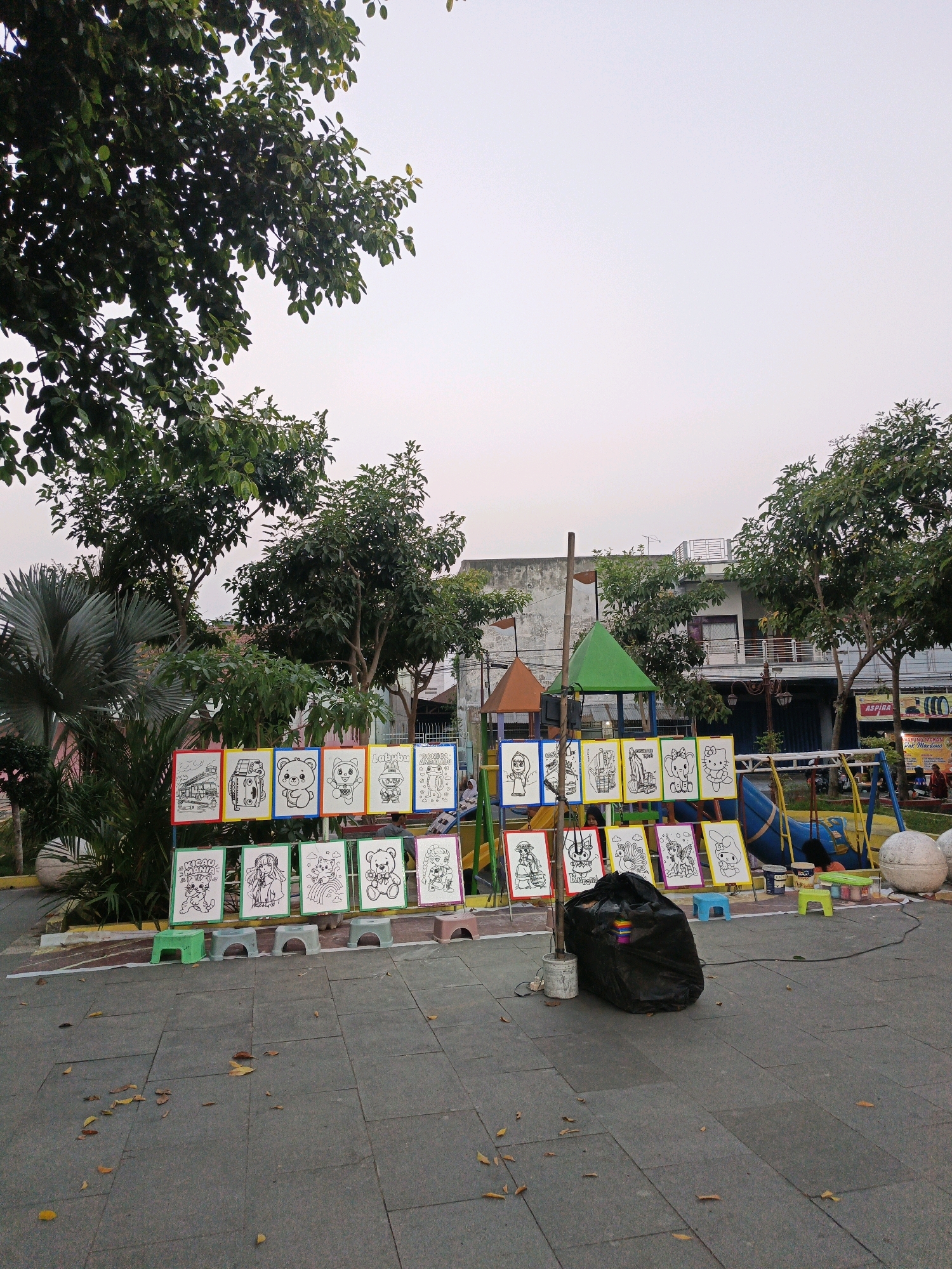 In Klaten, Indonesia, this image captures an outdoor scene featuring a playground area with several elements. In the foreground, a patterned paved area shows scattered fallen leaves. Rows of framed pictures, predominantly featuring cartoonish drawings, are displayed on easels. These drawings appear to be for children, with subjects like teddy bears, cars, and anime characters. Some of the pictures are still black and white outlines, suggesting they are for coloring.
Behind the display of drawings, a playground structure is visible, including a slide and swings. Various colorful plastic stools are placed around the display area. A large black trash bag is centrally located in front of some of the drawings, next to a white bucket.
Further back, buildings with balconies and windows are seen, along with trees and foliage. The sky is overcast. The overall impression is of a public space, possibly a park or recreational area, set up for children's activities, with the drawings serving as a coloring or art station. There are a few people in the background, some near the playground and others in the distance.