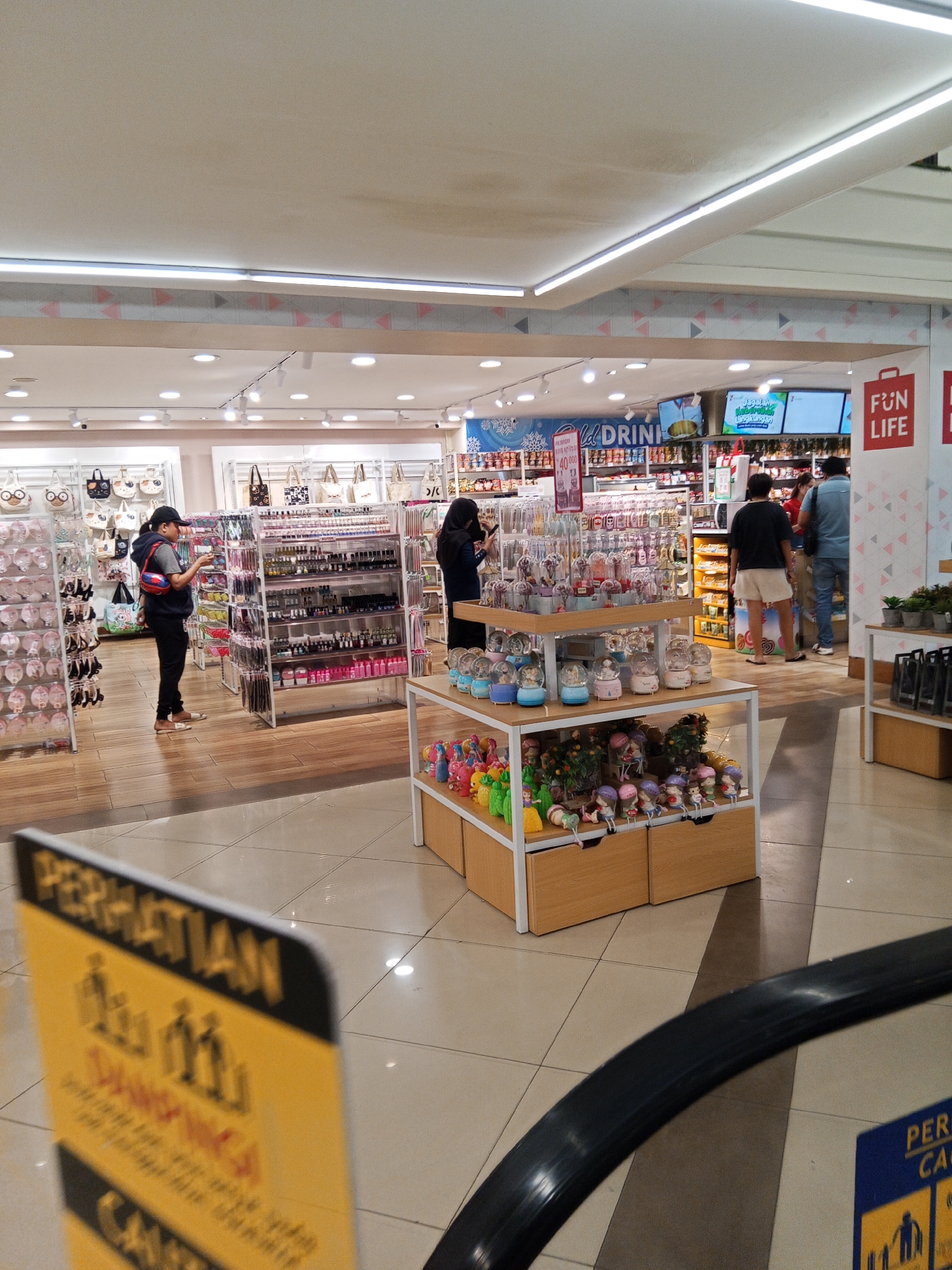 The image depicts the interior of a retail store, likely a department store or a specialty shop, potentially located in Lubuklinggau, Indonesia. The store features shelves stocked with various merchandise, including bags, cosmetics, and decorative items like snow globes and figurines. The flooring appears to be tiled, and the lighting suggests an indoor commercial setting.
In the foreground, a partially visible yellow sign with black text, likely a warning or informational sign, is present. In the mid-ground, several individuals are browsing or shopping. One woman, wearing a black jacket and a baseball cap, is examining items on a shelf. Another woman, in a dark top, is also looking at merchandise. A man in a light blue shirt and shorts is seen walking away from the camera.
The store's layout includes displays of merchandise on shelves and tiered stands. Signs with text in what appears to be Indonesian are visible throughout the store. One large sign above the shelves reads "DRINK". Another section of the store has signs that say "FUN LIFE". The overall atmosphere is that of a busy retail environment.
