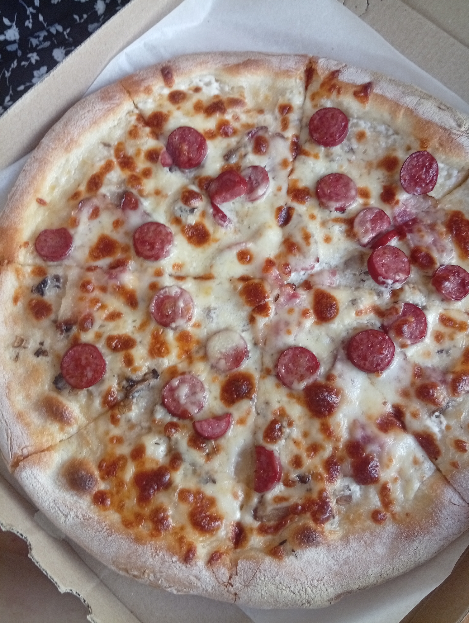 A whole pizza with sliced sausages and cheese is displayed in a cardboard pizza box. The pizza is cut into slices and is ready to be served. The background shows a patterned fabric and a white paper liner.