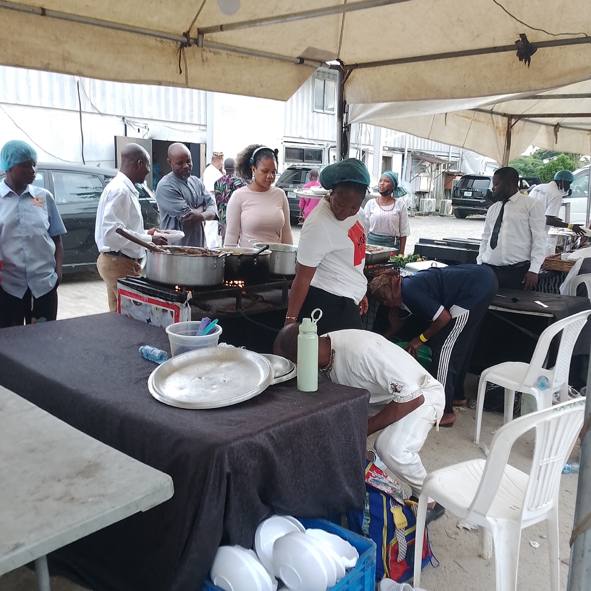 In Ikoyi, Nigeria, a bustling outdoor food stall operates under a beige tent. Several individuals are engaged in food preparation and service. A woman wearing a blue hairnet, a light blue collared shirt, and dark pants stands on the left, facing the food preparation area. Next to her, a man in a white shirt and tan pants is serving food from a large pot with a wooden spoon. Behind him, a man in a gray garment has his arms crossed. A woman in a pink long-sleeved shirt and a headscarf looks down at something near the food station. Another woman, wearing a white t-shirt with red text and a green headscarf, is also near the food preparation. To the right, a man in a white shirt and black tie is standing. A person in a blue uniform with white stripes on the pants is bending down near a table covered with a dark cloth. On this table, several large metal plates and a light green water bottle are visible. A blue plastic crate filled with white bowls is situated on the ground to the right of the table. White plastic chairs are placed around the seating area. In the background, several vehicles, including a black Range Rover, and air conditioning units are present.