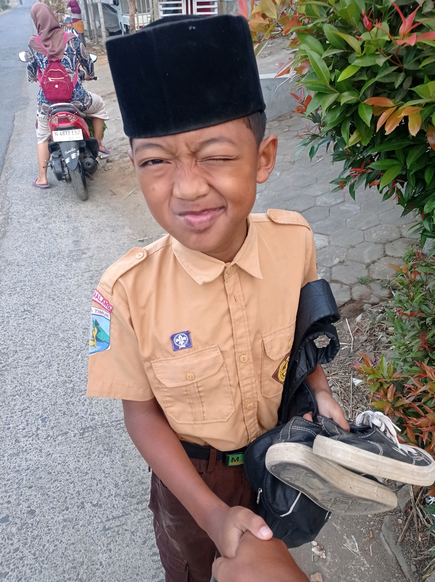 In the city of Purwosekar, Indonesia, a young boy, dressed in a tan school uniform with a black cap, playfully winks at the camera. He holds a black backpack and a pair of worn black sneakers in his hands. The boy's uniform features patches on the left sleeve, one depicting a landscape and text that appears to say "MALANG TIMUR," and a smaller blue scout emblem. Behind him, a woman on a red motorcycle, wearing a hijab and a patterned shirt, rides away down a paved street. The scene is set against a backdrop of foliage and a grey, patterned sidewalk. The lighting suggests it is daytime.