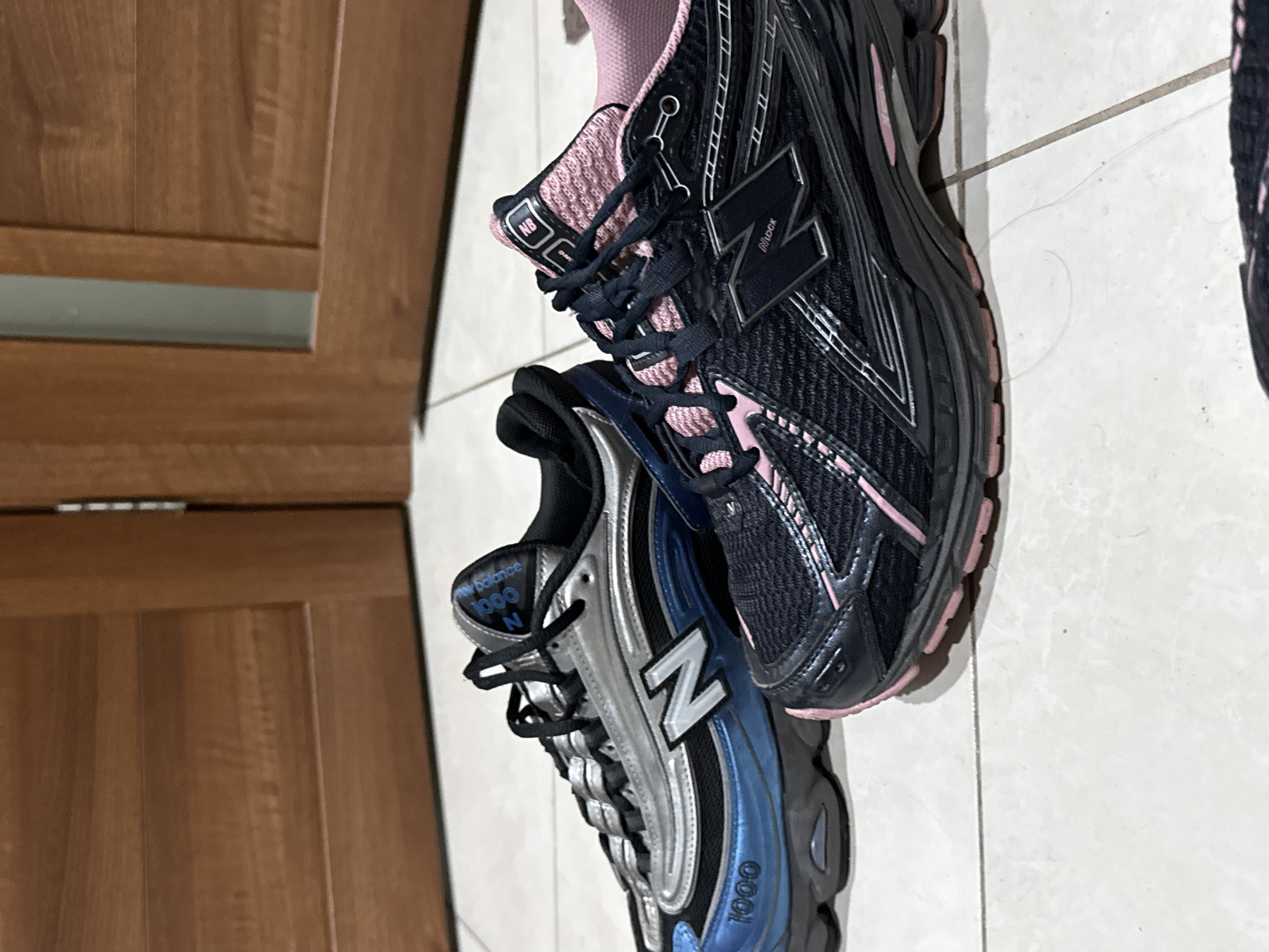 Two New Balance sneakers are displayed on a tiled floor in front of wooden doors. The shoe on the left is silver and blue with "1000" prominently displayed on the side and "N" logo on the side. The shoe on the right is black and pink with "NB" on the tongue and an "N" logo on the side. The scene is static and well-lit.