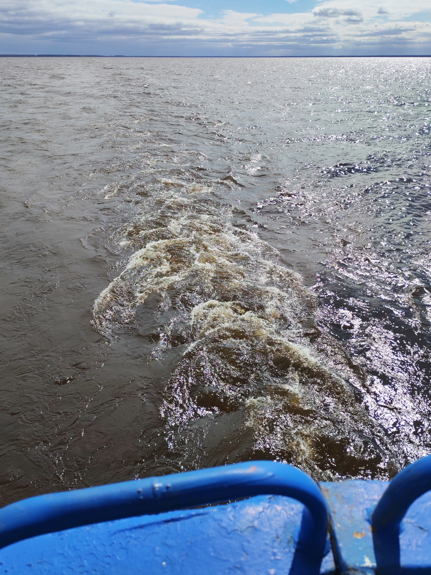 The image captures a view from a moving boat across a wide expanse of water under a partially cloudy sky. In the foreground, a weathered, bright blue metal railing is visible, indicating the perspective is from the vessel itself, looking out over the water.
The main subject is the water, which appears turbulent and murky, especially in the immediate wake of the boat. A prominent, foamy trail of churned water, brownish in color with sparkling highlights from the sun, extends from the lower center towards the mid-right of the frame. To the left of the wake, the water is a darker, more uniform brown. To the right, and further out, the water reflects intense sunlight, creating numerous bright, shimmering points across its surface. The overall impression of the water is choppy and restless.
In the mid-ground, the vastness of the water continues, stretching towards a distant, low-lying horizon line. A faint, dark strip of land or shoreline can be seen along the horizon. Above this, the sky occupies the upper portion of the image, featuring a mix of white and grey clouds against a lighter blue backdrop, suggesting a partly sunny day. The sunlight is strong enough to create significant glare on the water.
The scene depicts the activity of traveling by boat on a large body of water, likely a river or reservoir given the context of Mologa, Russia. The time of day is daytime, with clear indications of sunlight. There is no visible text in the image.