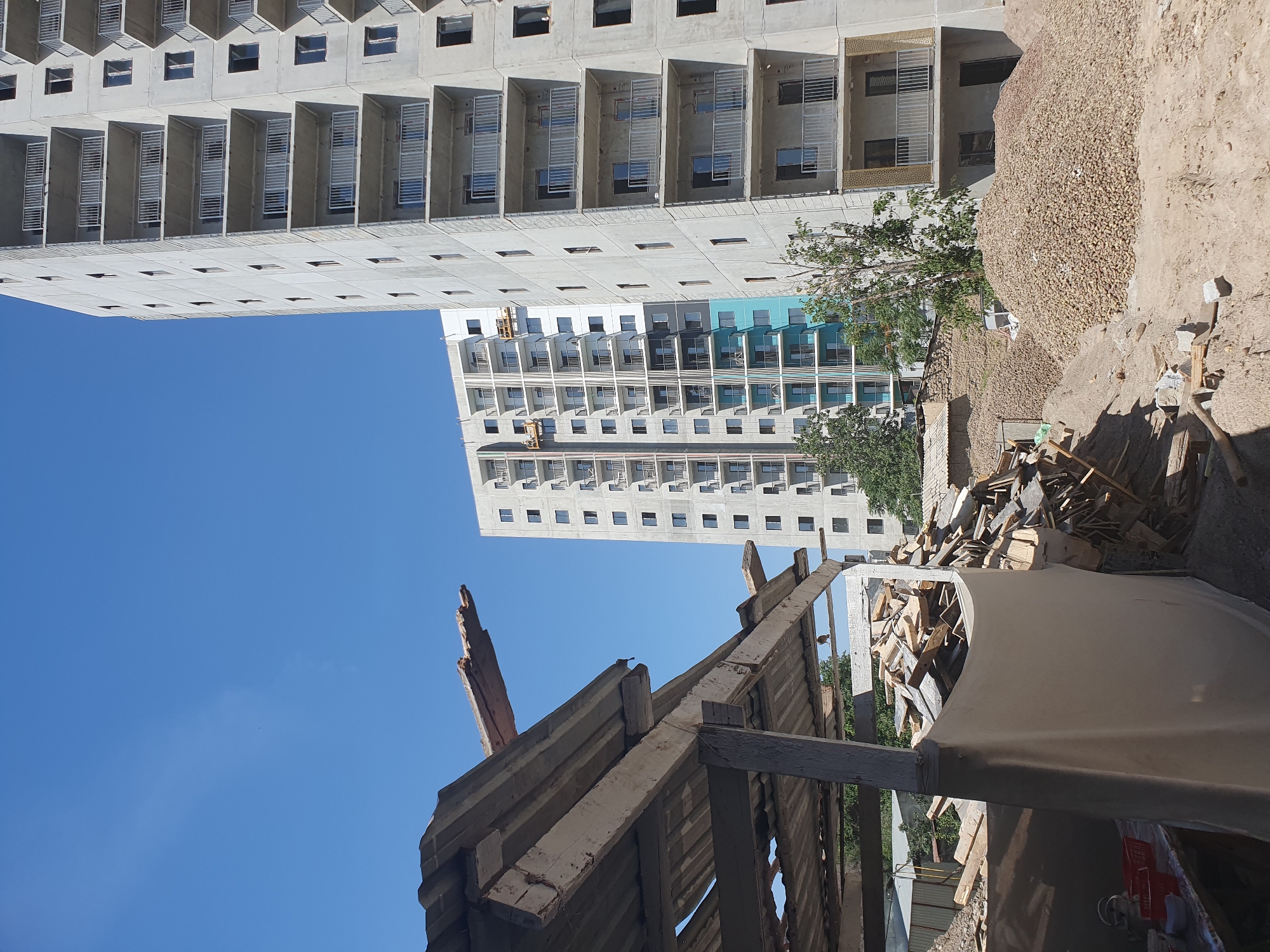 The image captures a construction site in Tashkent, Uzbekistan, under a clear blue sky. Two modern apartment buildings, still in various stages of construction, dominate the background. The building on the right is taller and appears closer to completion, featuring multiple balconies with metal railings and unfinished windows. The building to its left is slightly shorter and shows some exterior tiling in a turquoise blue. In the foreground, a dilapidated wooden structure with a corrugated metal roof is visible, suggesting an older part of the site or demolition work in progress. Piles of building debris, including wood scraps and what appear to be potatoes, are scattered across the sandy ground. The overall scene depicts a dynamic urban development project.