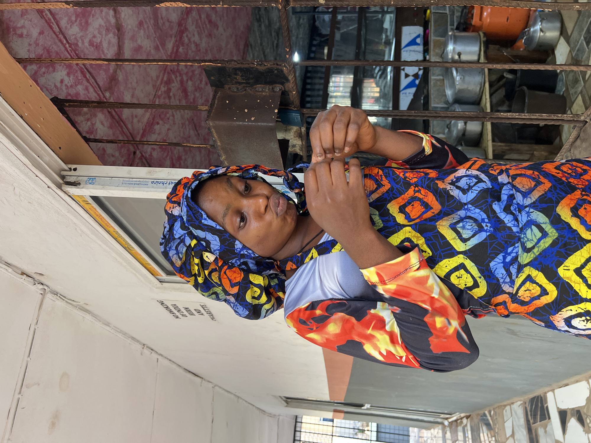 The image shows a young Black woman in Nkpor, Nigeria, wearing a colorful headwrap and a brightly patterned dress. The dress features a vibrant design with squares in blue, orange, yellow, and white, and a sleeve with a fiery, flame-like pattern. She is positioned looking towards the camera, with her hands clasped in front of her chest. Her expression is neutral, with her lips pursed slightly.
Behind her, a metal grate is visible, suggesting a window or barrier. Beyond the grate, shelves are laden with various pots and cooking utensils, including a large orange gas cylinder. The setting appears to be an indoor or semi-enclosed space, possibly a shop or a storage area. A white wall with some staining is present to the left of the woman, and a sign with text is partially visible on it.
The text on the wall reads, "NO SMOKING AND BEEN OF CIGARETTE". The lighting suggests it could be daytime. The overall impression is a candid shot of a person in a functional, perhaps commercial, environment in Nigeria.