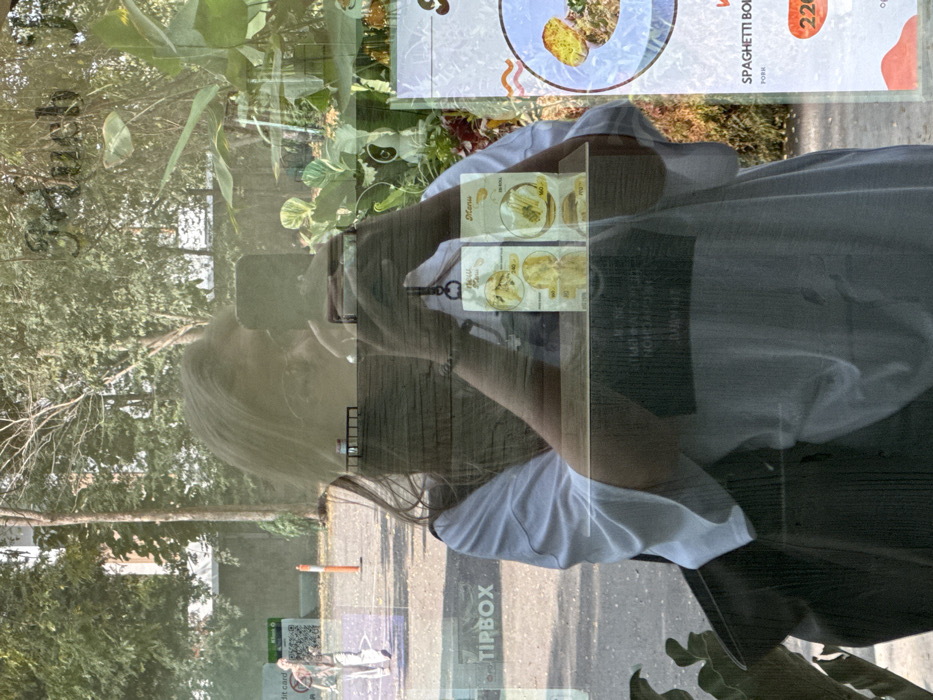 A reflection in a window shows a woman taking a photo with her phone. The reflection is overlaid with the restaurant's menu displaying pasta dishes. Outside, a street with trees and a Tipbox advertisement is visible. The woman wears a white t-shirt, and her hair is blonde. The restaurant's interior is hinted at with leafy plants.