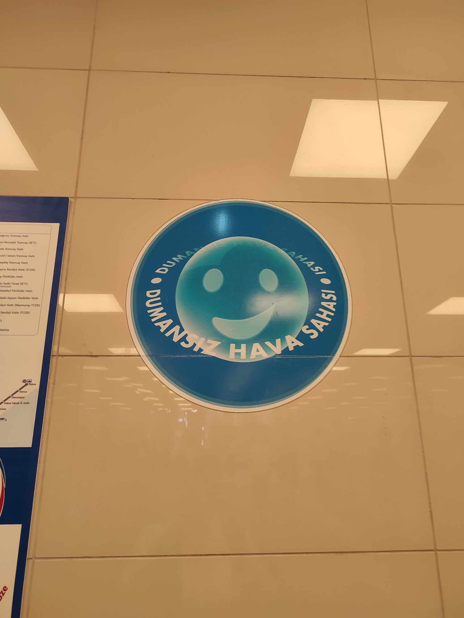 The image shows a circular blue and white sticker with a smiley face on a tiled wall. The sticker reads "DUMANSIZ HAVA SAHASI" in Turkish, which translates to "Smokefree Air Area." The wall is made of light brown tiles with visible grout lines and illuminated by overhead lights which cast reflections. To the left of the sticker, a portion of a blue and white informational poster is visible, showing what appears to be a map or diagram with text in Turkish. The text on the poster includes place names and route information, suggesting it might be related to public transportation or local amenities. While no people are visible in the frame, the presence of the informational poster and the public space setting suggest this is a public area such as a metro station, bus terminal, or a public building. Given that Maltepe is a district in Istanbul, Turkey, this sticker and poster are likely located within Maltepe.