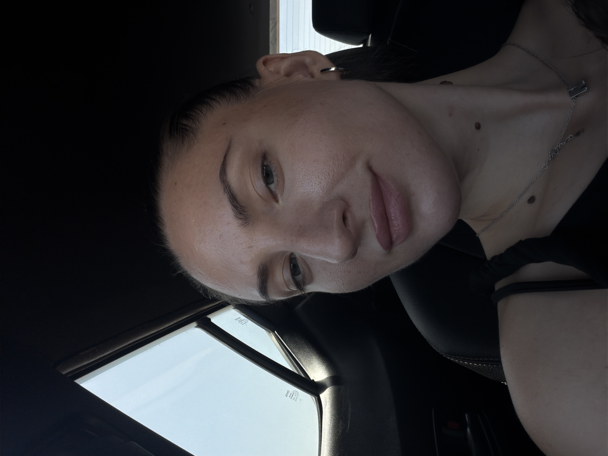 A selfie is taken from the chest up of a woman in a car, looking forward. She has her hair pulled back and is wearing a black tank top and a silver necklace. A car window is visible behind her, showing a bright sky. There are several moles on her neck. The overall lighting suggests it is daytime. The image location in Sharm El Sheikh, Egypt is not discernible from the visual content.