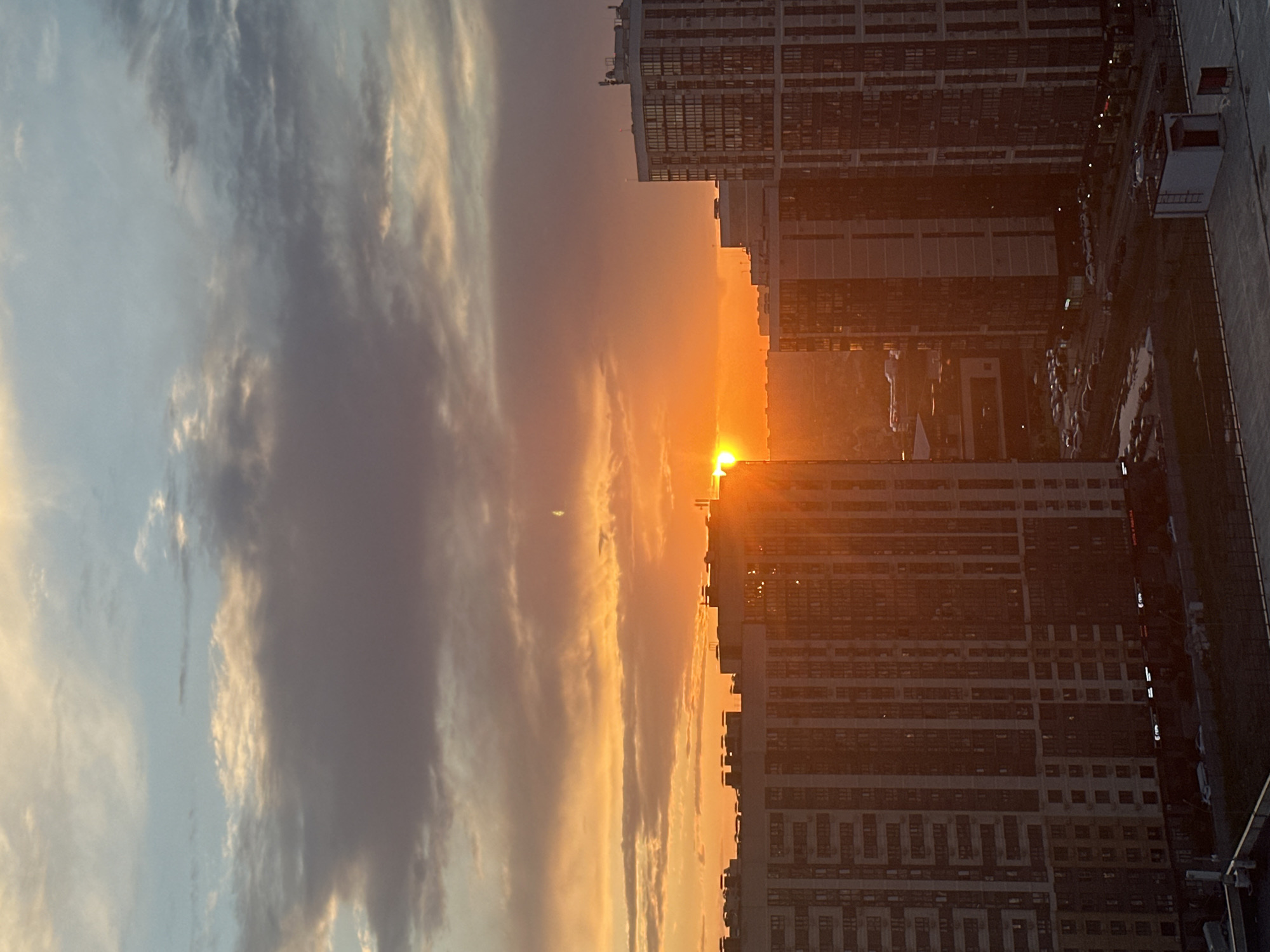 The image depicts a panoramic view of a city skyline at sunset, likely in Krasnodar, Russia. Tall residential buildings with numerous windows dominate the foreground and midground, their facades illuminated by the warm, orange glow of the setting sun. The sky above is a canvas of dramatic clouds, transitioning from a pale blue near the top to vibrant shades of orange, pink, and grey closer to the horizon where the sun is partially obscured by buildings. The sun itself appears as a bright, concentrated point of light casting long shadows. Below the buildings, a street level scene is visible with parked cars and a few scattered structures. The overall atmosphere is one of urban development bathed in the dramatic light of sunset.