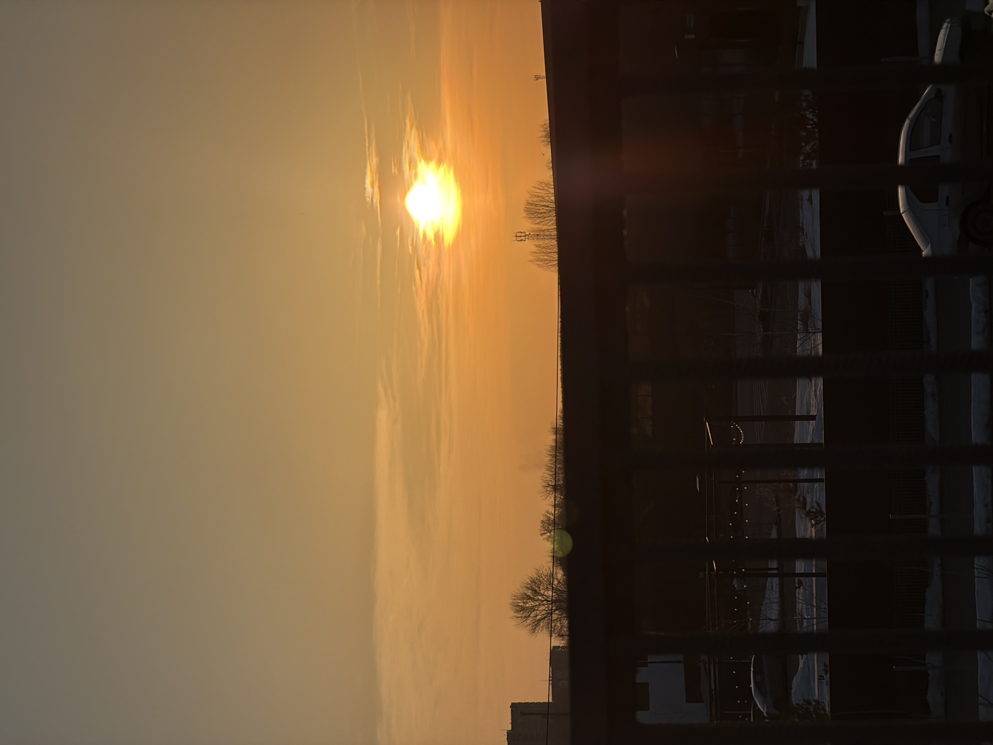 The scene depicts an urban or suburban landscape in Izhevsk, Russia, viewed during either sunrise or sunset. The sky is predominantly clear with an orange-yellow hue, featuring a bright, intense sun positioned low on the horizon, partially obscured by light clouds.
In the immediate foreground, a dark, vertical-bar barrier, resembling a fence or balcony railing, partially obstructs the lower half of the frame. Below this barrier, a dark horizontal structure is visible.
The midground reveals ground patches covered in snow, indicating a cold season. A light-colored sedan, possibly a Renault Logan, is visible in profile, parked or moving slowly on a dark surface, likely a street. Another darker vehicle is partially visible further to the left. Bare trees are scattered throughout this section. Artificial lighting elements, including string lights, are visible, suggesting an illuminated public or commercial space.
The background consists of a silhouette of bare deciduous trees and multiple communication towers, including a prominent lattice structure located centrally on the horizon and another smaller tower to its right. Distant buildings, including a multi-story structure on the far left, are also silhouetted against the bright sky. No individuals or specific actions are discernible within the image.