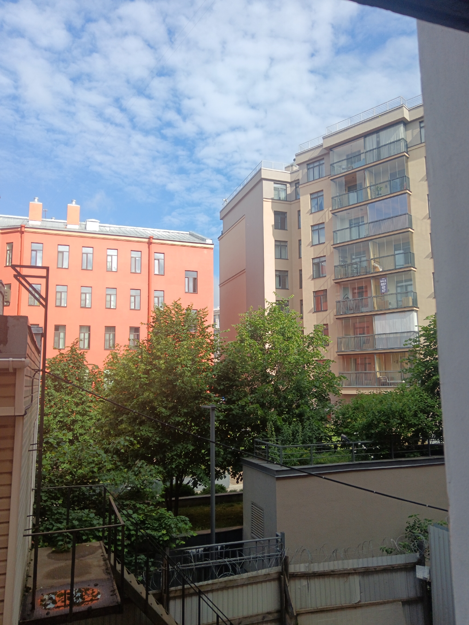 The image shows a view of apartment buildings and trees under a bright blue sky with scattered clouds. In the foreground on the left, a beige-colored building with horizontal siding is partially visible, along with an external staircase made of metal. The staircase has a wet surface with a graffiti-like marking on it.
Behind this structure, a bright orange building with many windows spans across the mid-left of the frame. Two chimney-like structures are visible on its roof. To the right of the orange building, a taller, light beige apartment building with glass-enclosed balconies dominates the right side of the image. A sign with the text "SALE 627 08 67" is attached to one of the balconies.
Dense green trees with lush foliage are positioned between the buildings, with their branches reaching upwards. In the background, the sky is a clear blue, dotted with fluffy white clouds, suggesting a pleasant daytime with fair weather. The overall scene depicts an urban or suburban residential area.