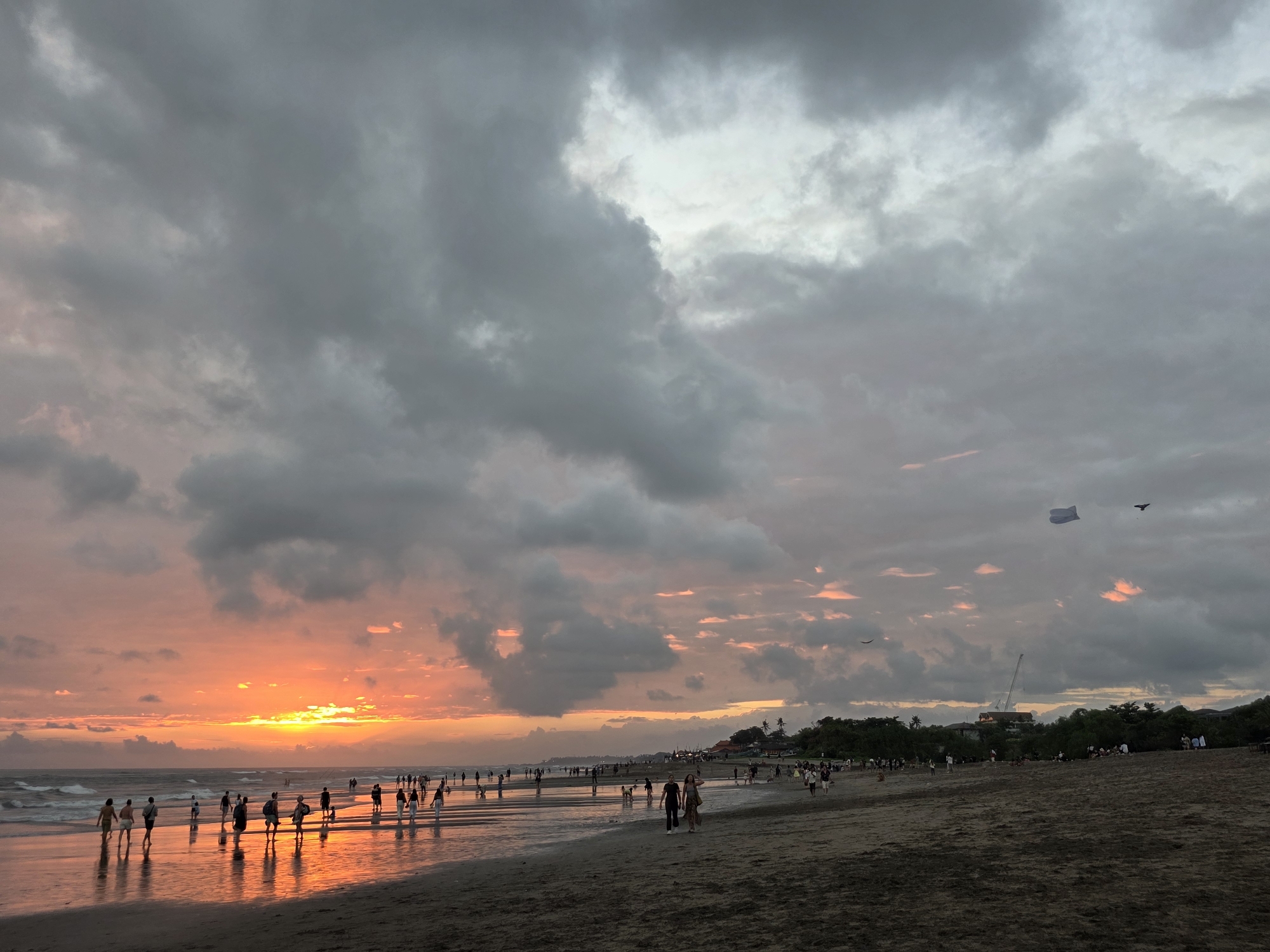 The image depicts a beach scene in North Kuta, Indonesia, during what appears to be sunset. The sky is heavily overcast with dark, dramatic clouds, interspersed with patches of vibrant orange and pink hues, indicative of the setting sun. The ocean in the foreground exhibits moderate waves.
Numerous people are scattered across the sandy beach and the shallow water. Some are walking along the shoreline, while others appear to be standing or interacting in small groups. Many have their reflections mirrored on the wet sand. In the background, a line of trees and some structures can be observed along the edge of the beach. Two kites are visible in the sky, one white and one dark, along with what appears to be a bird. The overall atmosphere is one of a busy public beach during twilight.