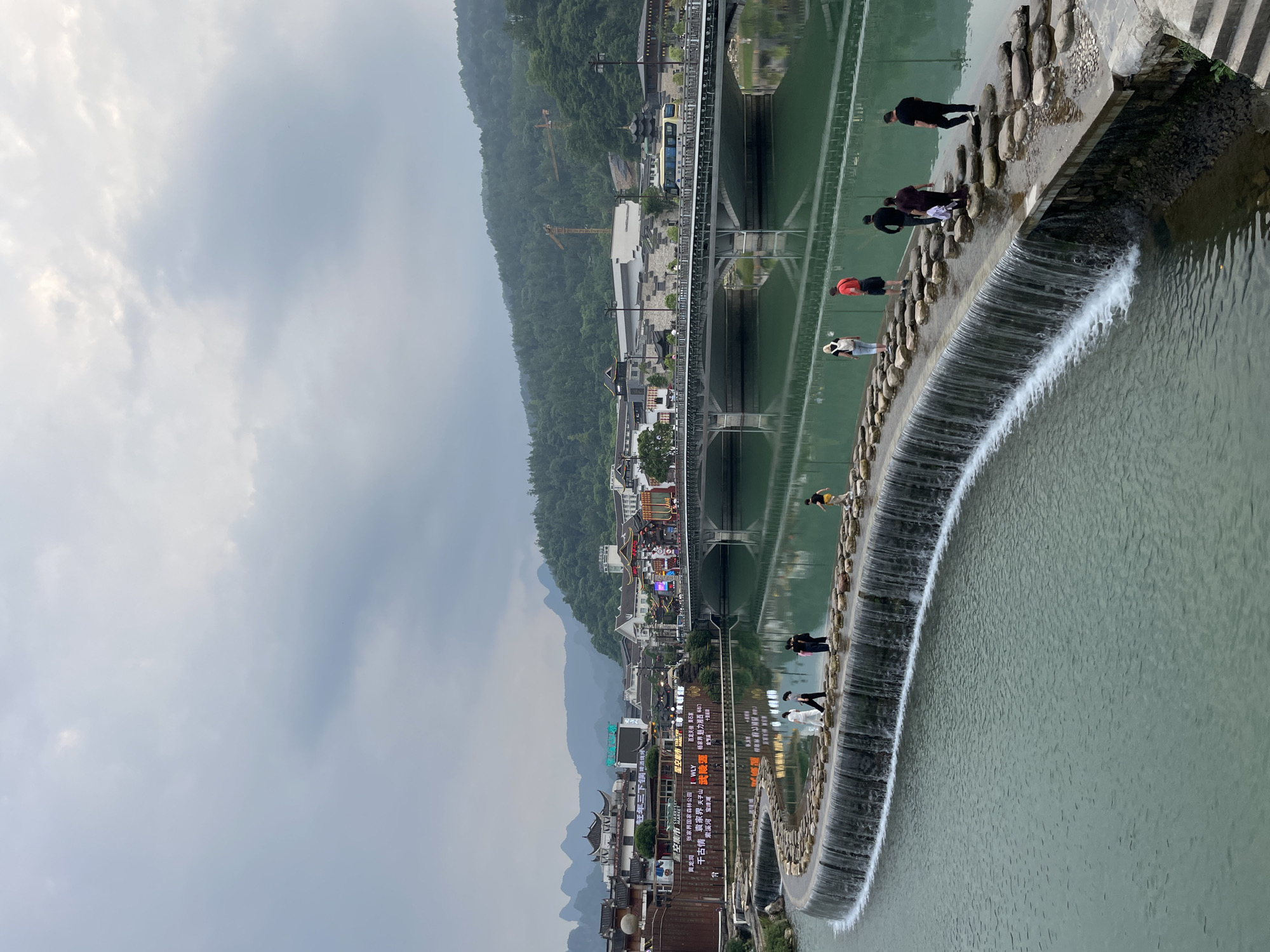 The image captures a panoramic view of Wulingyuan, China, showcasing its unique topography and architectural elements. In the foreground, a constructed circular dam creates a waterfall effect. People are observed walking on this structure. The river reflects the buildings and bridges above. The urban area extends into the distance. Hills covered in verdant trees frame the backdrop, with a cloudy sky overhead. Text on signs is visible, including the phrase "Zhangjiajie National Forest Park."