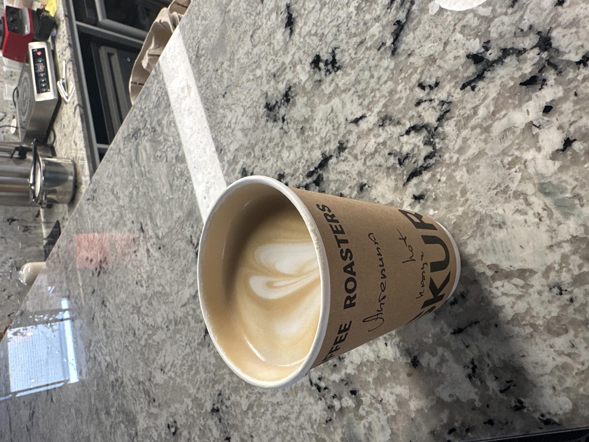A cup of coffee sits on a gray countertop with black and white speckled pattern. The cup has the words "COFFEE ROASTERS" and "Amrenuna" handwritten on it. Also on the cup is "KUU", and "Kanzur hot". It appears to be latte art on top. Behind the cup is a counter with appliances including a stainless steel pot and electric stove top. It is not possible to determine the location of the city of Kazan, Russia from the provided image.