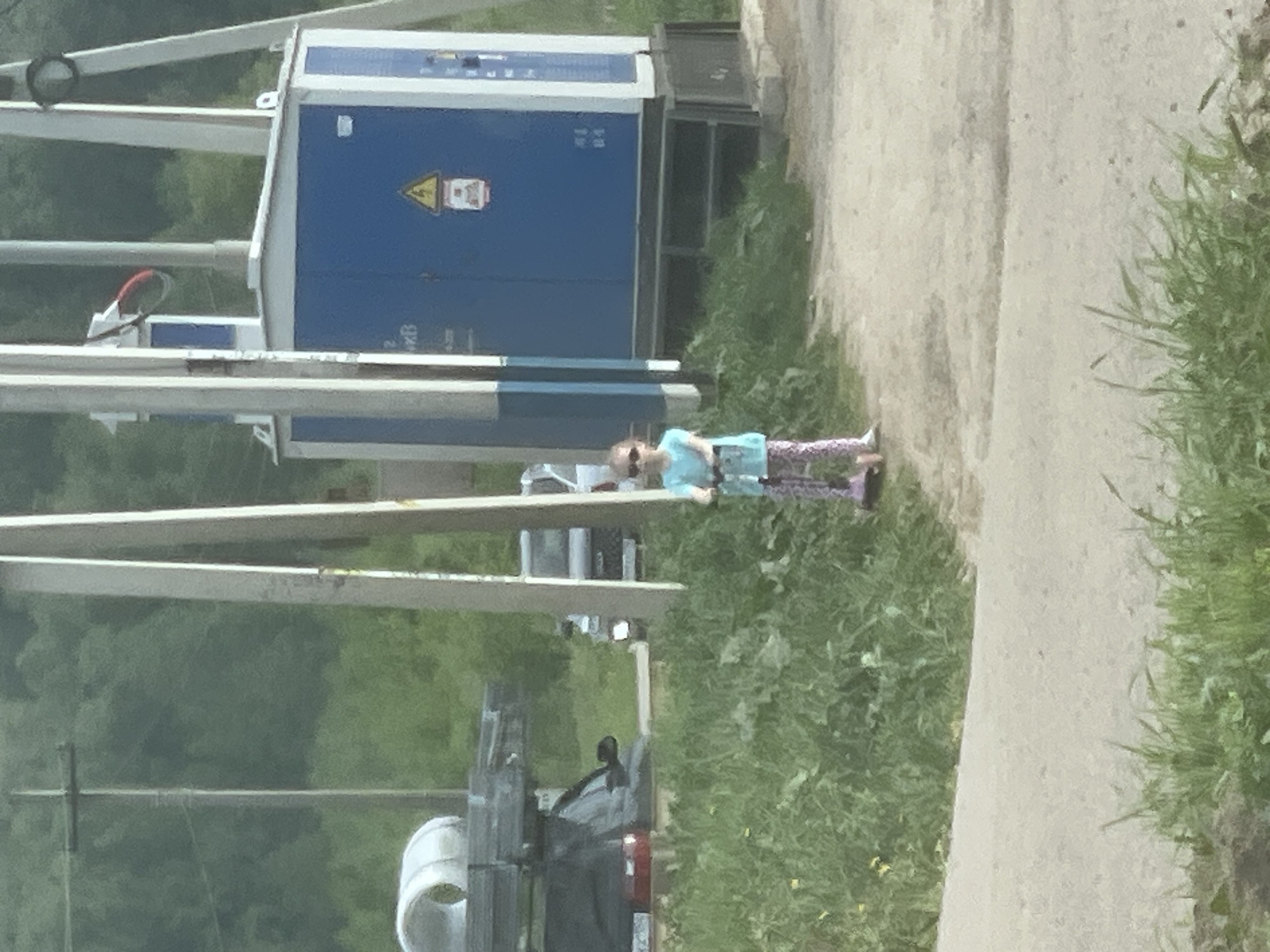 The image depicts a scene in Molokovo, Russia. A child, wearing a light blue shirt, patterned leggings, and purple shoes, stands on a grassy area alongside a paved path. Behind the child is a gray vehicle with a white container on its rear. To the left of the vehicle are power lines and a blue electrical transformer with warning symbols, indicative of a rural or industrial area. The background shows green trees, suggesting it's either a park or the outskirts of the city.