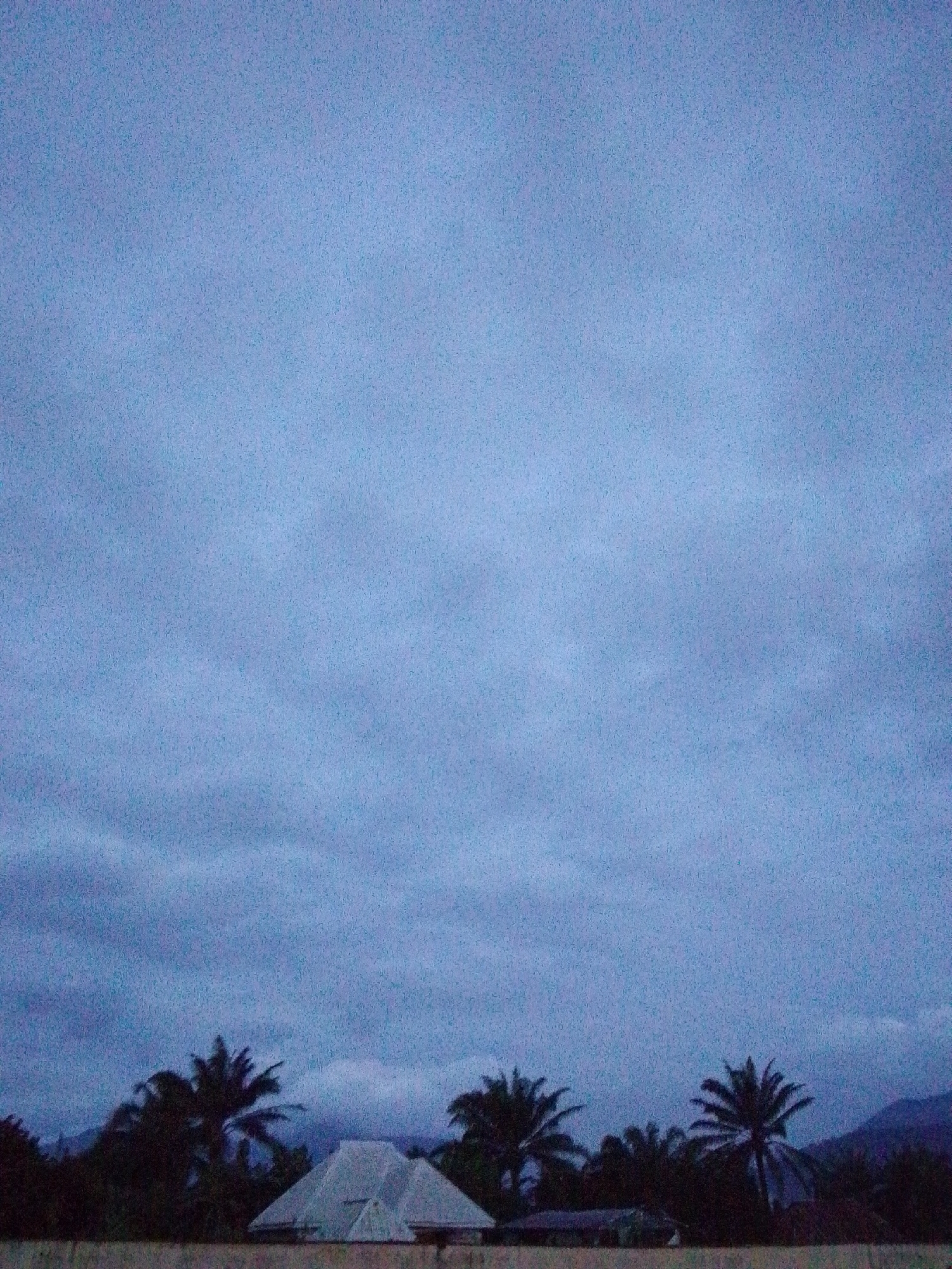 The image depicts a cloudy, twilight sky over a rural landscape. In the foreground, a house with a light-colored, peaked roof is visible, partially obscured by silhouetted palm trees and other foliage. The scene appears to be an outdoor view, likely from ground level, with a clear horizon line where the sky meets the land. The overall lighting suggests it is either dawn or dusk. The location is identified as Serti, Nigeria.