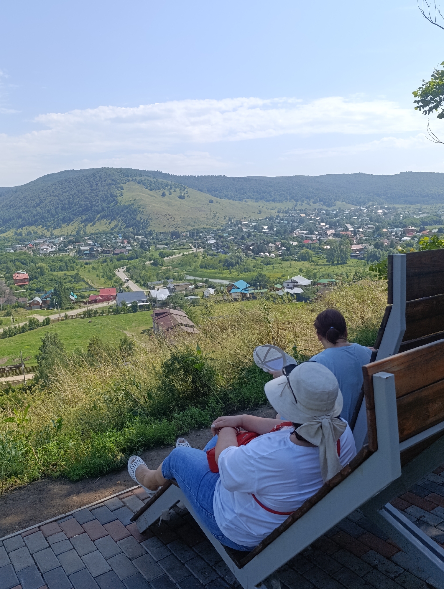 Two individuals are seated on a modern outdoor bench, positioned on a paved area overlooking a sprawling valley. The person in the foreground, wearing a white t-shirt, blue jeans, and a wide-brimmed hat tied with a beige scarf, is facing away from the camera. Their right hand rests on their leg, and their feet are clad in white sandals. Beside them, a second individual, seen from behind, wears a blue t-shirt and has their hair tied back in a ponytail. They are holding a light-colored cap. The bench features wooden slats and a metallic frame.
The background reveals a picturesque landscape with a village nestled in a valley. Numerous houses, characterized by red and grey roofs, are scattered amongst lush green fields and trees. A road winds through the settlement, with a white vehicle visible on it. In the distance, rolling hills covered in dense forest rise towards a bright blue sky dotted with scattered white clouds. The overall scene suggests a scenic viewpoint in the city of Solnechnaya Polyana, Russia.
