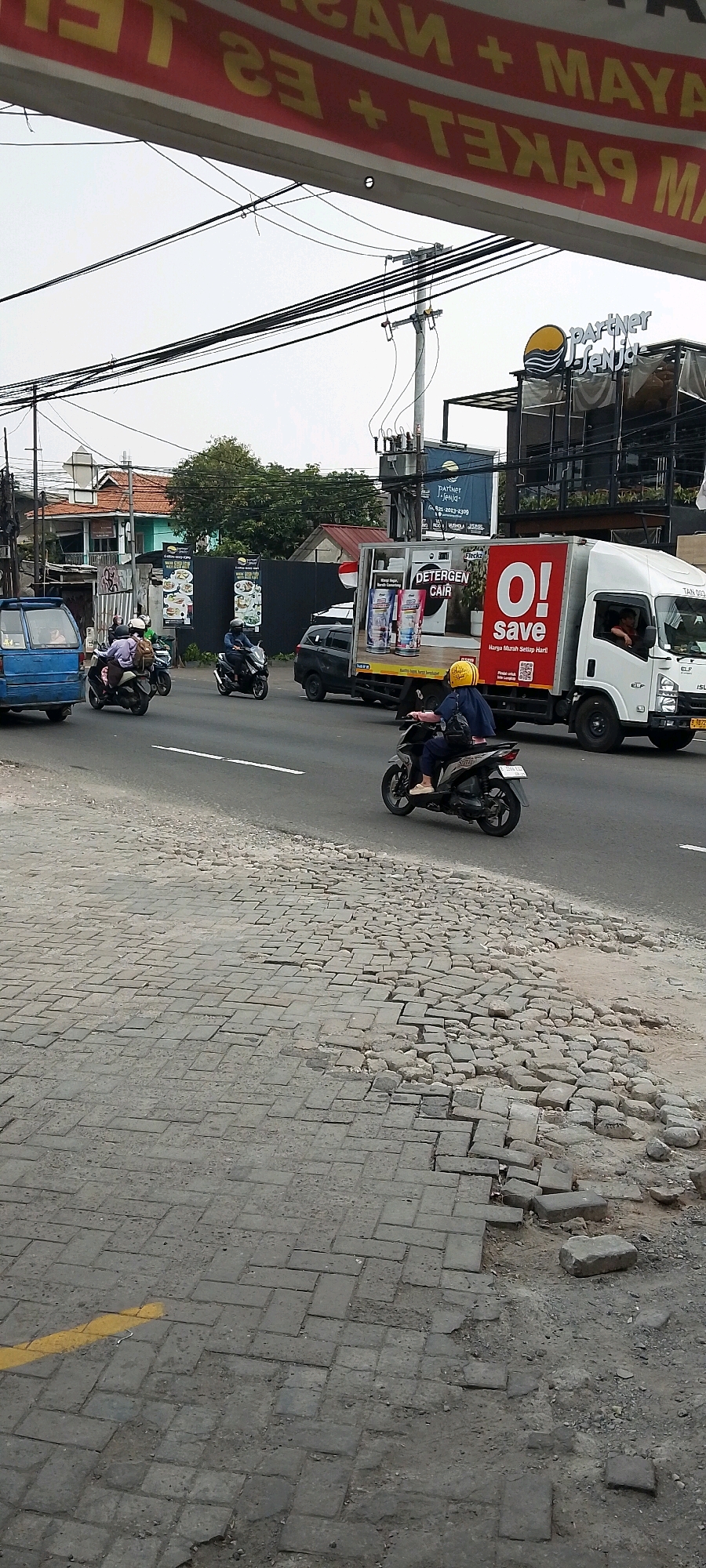 The image captures a street scene in Depok, Indonesia. The foreground shows a broken and uneven brick sidewalk with a yellow line painted on it. The street itself is paved and has several vehicles on it, including a blue pickup truck, several motorcycles, a black SUV, and a white truck. The white truck has a large advertisement on its side for "DETERGEN CAIR" and "O! save." One person is visible riding a motorcycle in the foreground with a yellow helmet. In the background, there are buildings, trees, utility poles with numerous wires, and a sign for "partner JENidq." The sky is overcast.