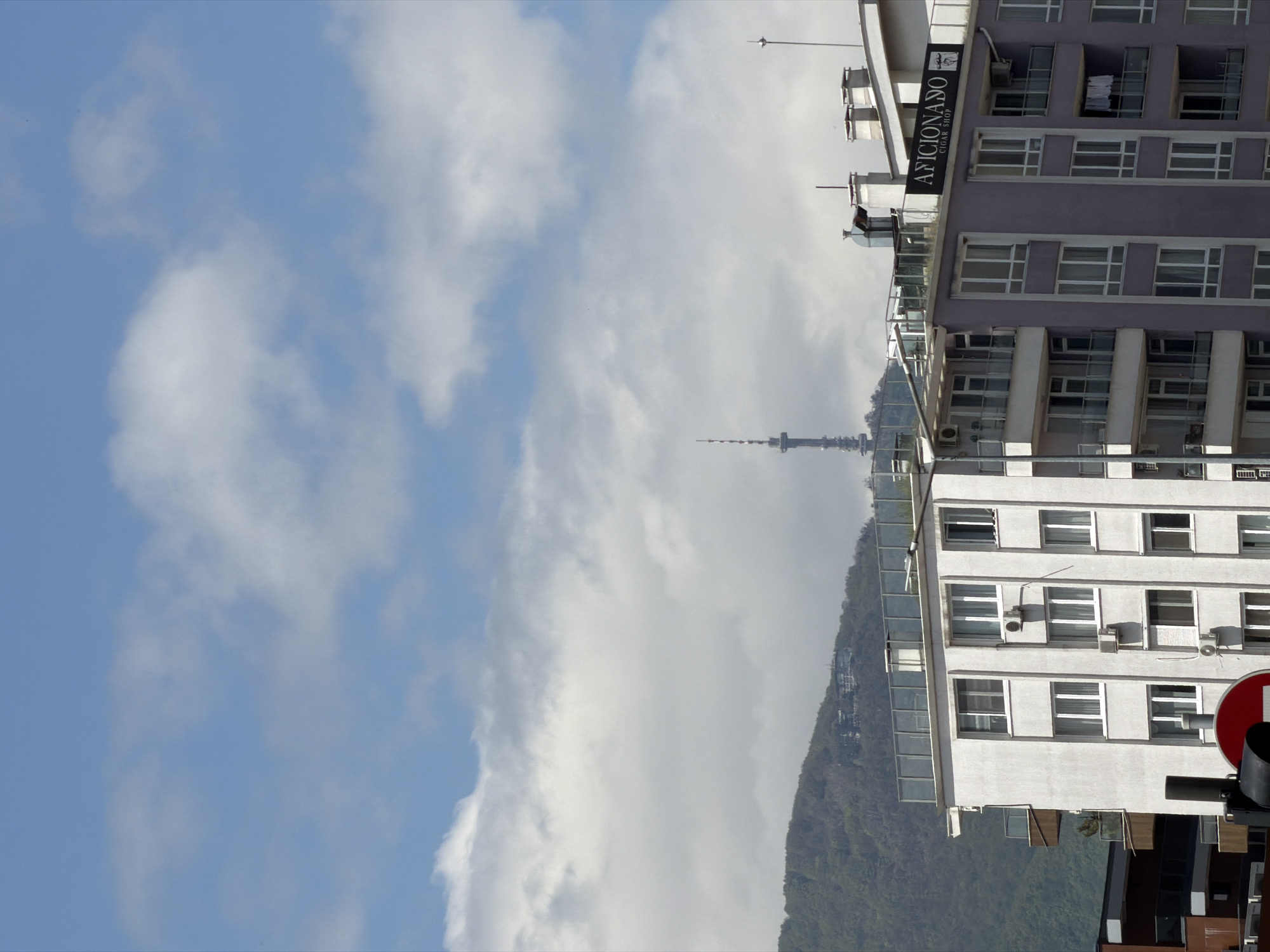 The image shows a partial view of Sofia, Bulgaria. The sky is visible, featuring a bright blue sky with white clouds. There are two buildings on the right side of the frame. One is white, while the other is painted purple. The sign on the purple building is for "Aficionado Cigar Shop". In the center of the image, there is a tower behind a green hill. It is most likely the TV Tower.